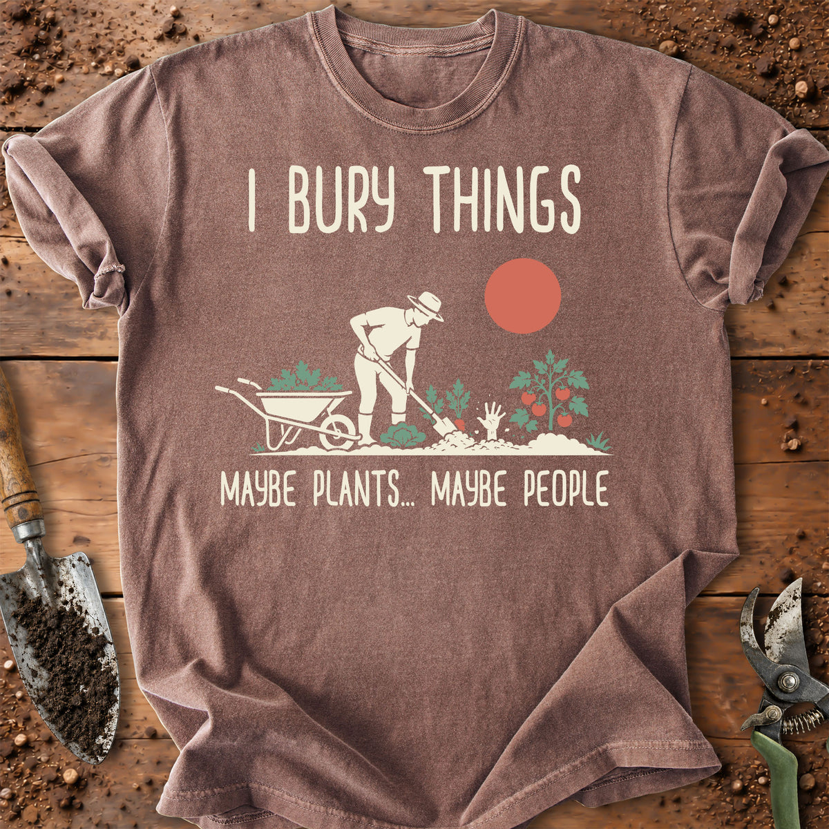 A brown graphic t-shirt laid flat on a rustic wooden surface with gardening props like a trowel and pruning shears. The shirt features a cream-colored graphic of a gardener burying a wheelbarrow with the text 'I BURY THINGS' above and 'MAYBE PLANTS.