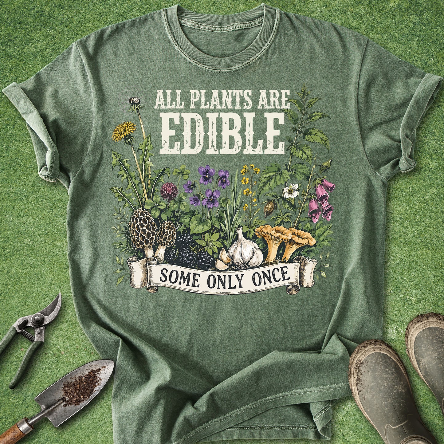 An olive green graphic t-shirt laid flat on a green grass-like surface, surrounded by gardening props including pruning shears, a trowel, and boots. The shirt features a graphic of various edible plants, flowers, garlic, and mushrooms with the text 'ALL PLANTS ARE EDIBLE' and 'SOME ONLY ONCE'.