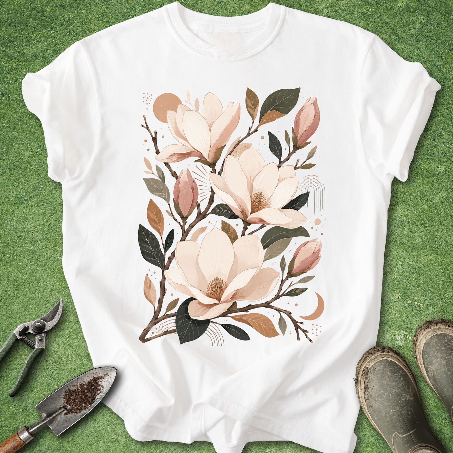 A white short-sleeve T-shirt laid flat on green grass, featuring a large floral print of magnolia blossoms in soft blush, beige, brown, and muted green tones. Gardening tools and muddy boots are placed around the shirt as styling props.