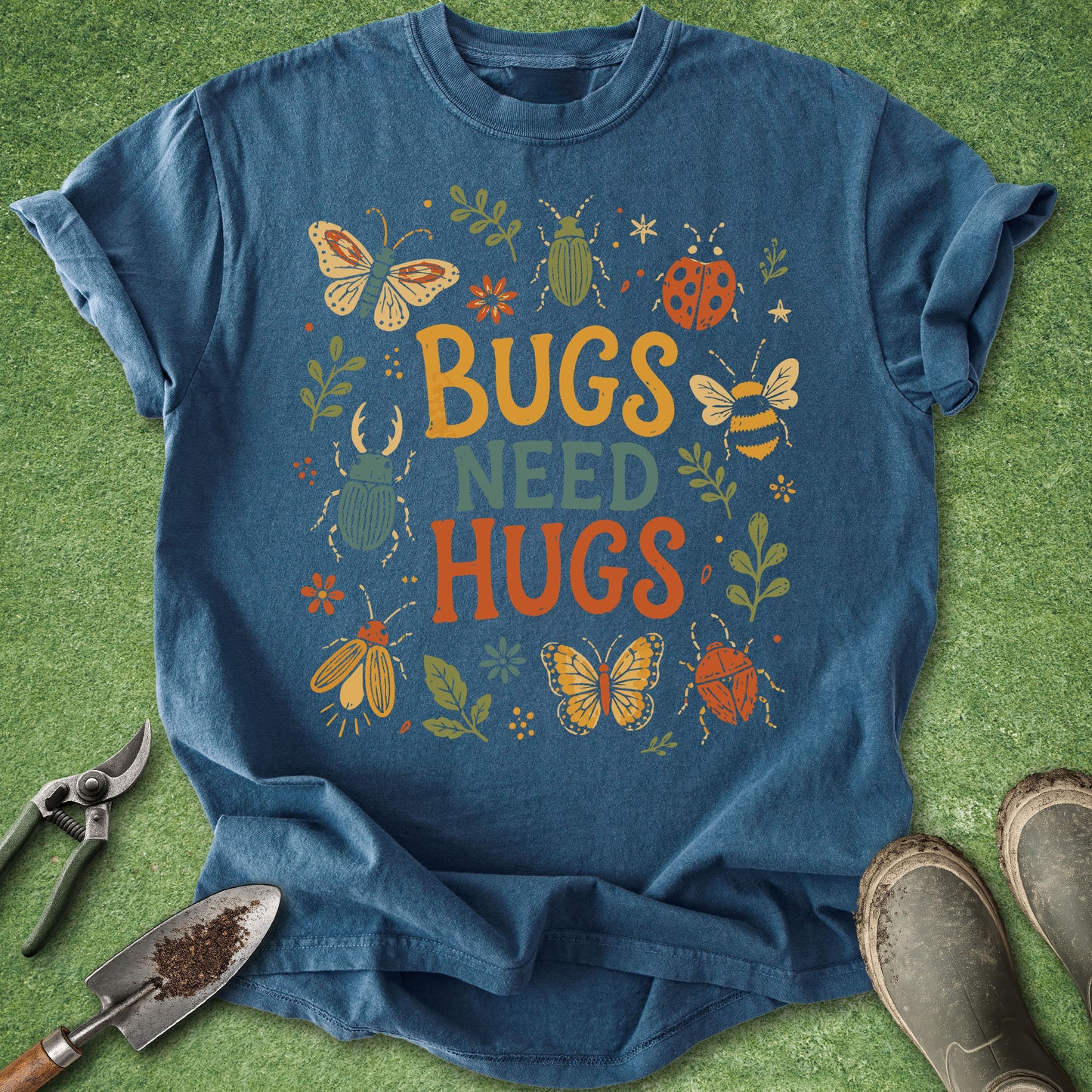 A blue graphic t-shirt laid flat on a green grass-like surface, surrounded by gardening props including pruning shears, a trowel, and boots. The shirt features a colorful design with the text 'BUGS NEED HUGS' surrounded by illustrated butterflies, bees, ladybugs, and leaves.
