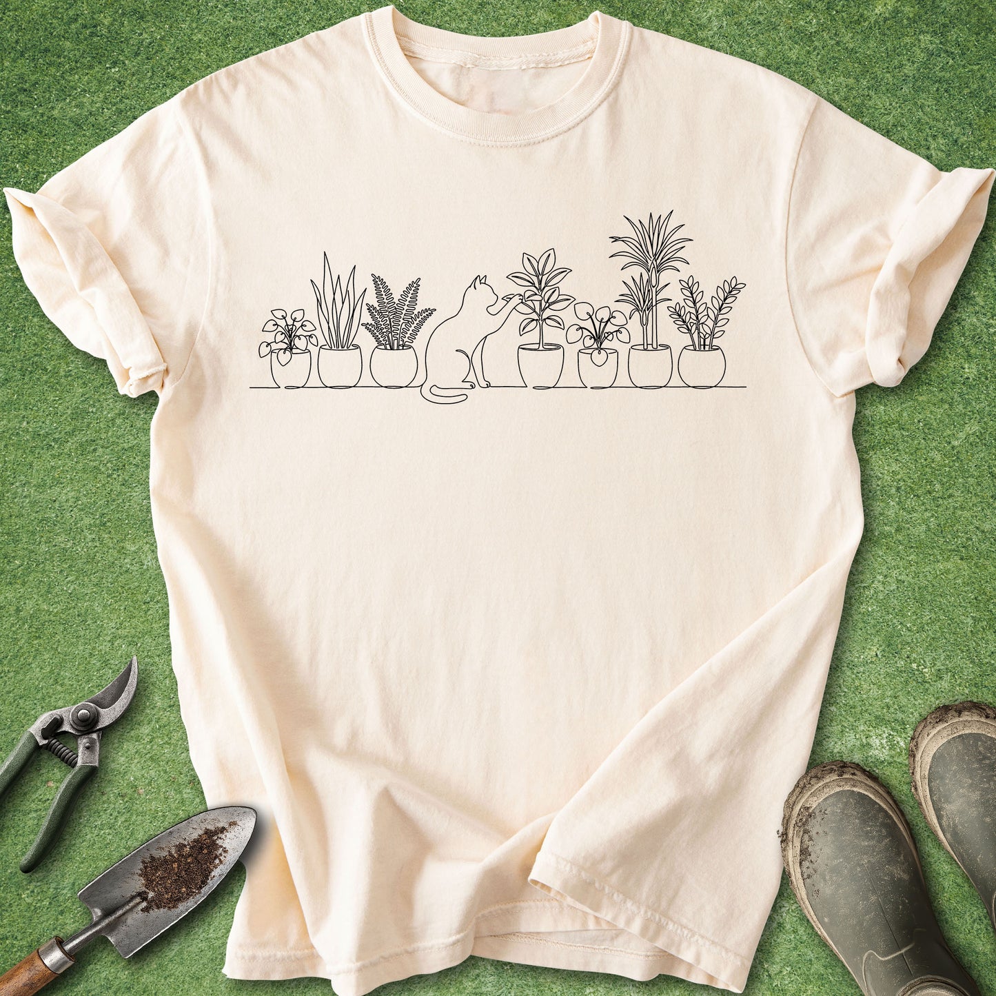 A cream-colored short-sleeve t-shirt with a black line art graphic of potted plants and a cat, laid flat on a green grass-like surface with gardening tools and boots nearby.