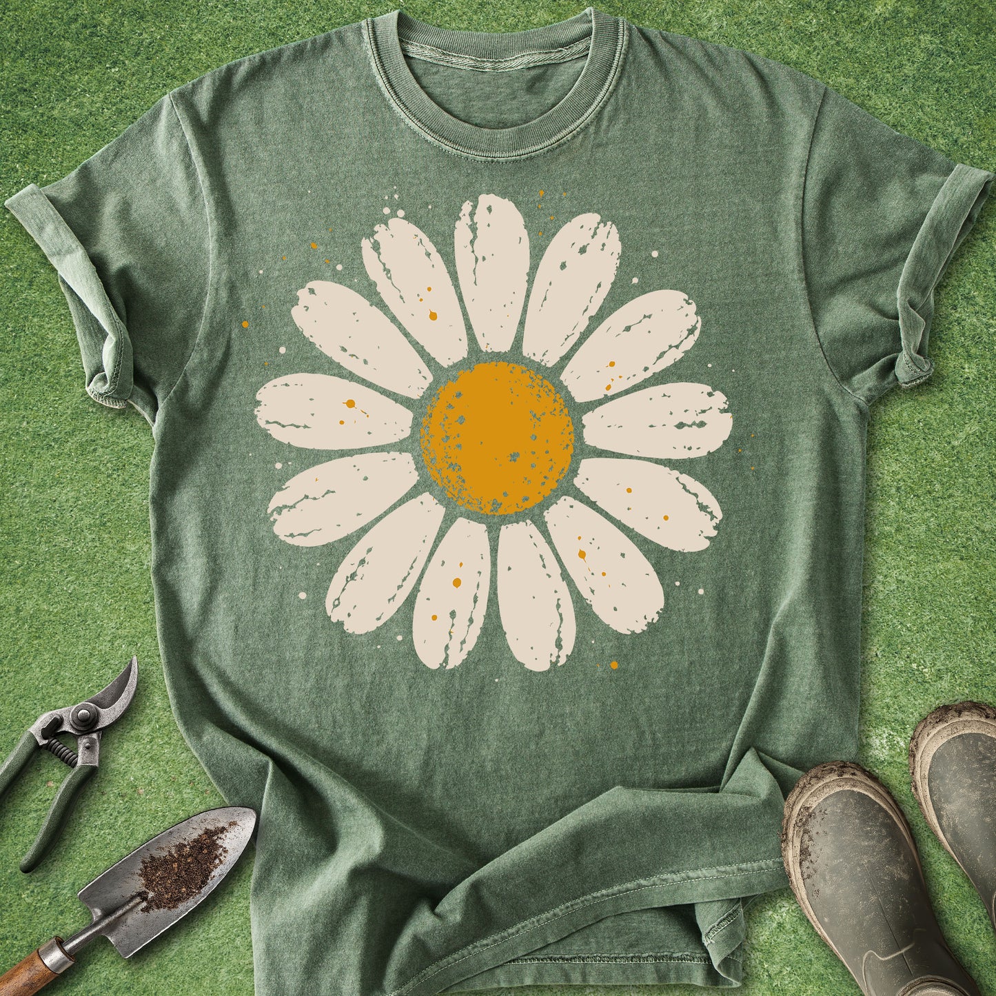 A green short-sleeve t-shirt with a large white daisy graphic featuring a yellow-orange center, laid flat on a green grass-like surface with gardening tools and boots nearby.