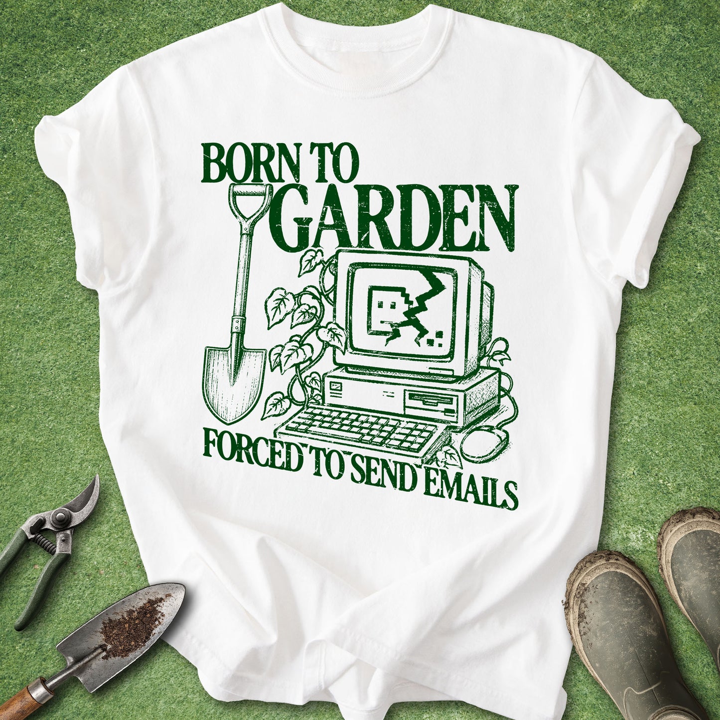 A white short-sleeve graphic T-shirt laid flat on green grass-like background, with a dark green printed design reading 'BORN TO GARDEN FORCED TO SEND EMAILS' around an illustration of a computer, shovel, plants, and tomatoes. Gardening props including pruning shears, trowel, seedling, and boots are placed around the shirt.