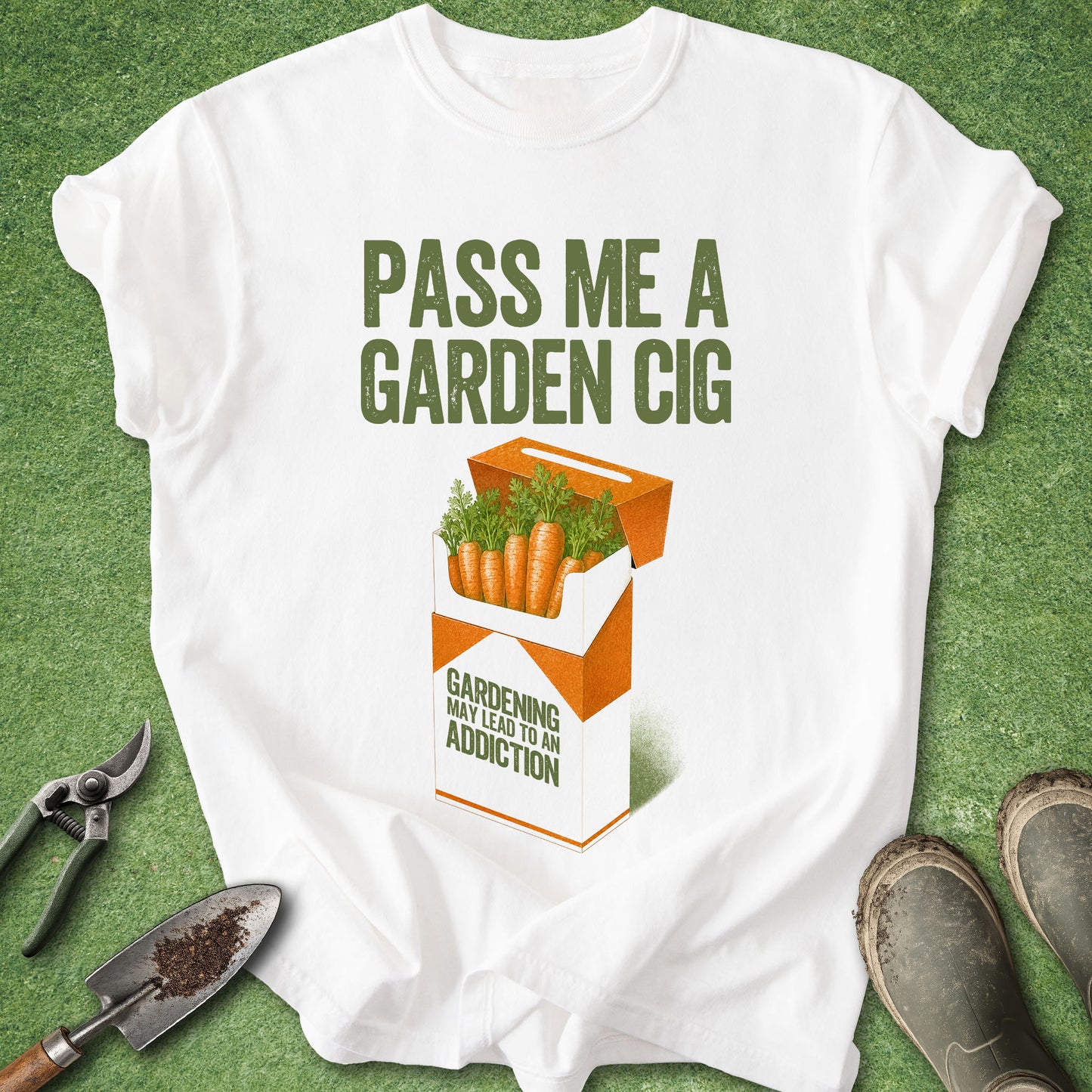 A white graphic t-shirt laid flat on green grass, featuring a humorous design that compares a garden bag to a cigarette pack with carrots and the text 'PASS ME A GARDEN CIG' and 'GARDENING MAY LEAD TO AN ADDICTION'. Gardening props like pruning shears, a trowel, and boots are arranged around the shirt.