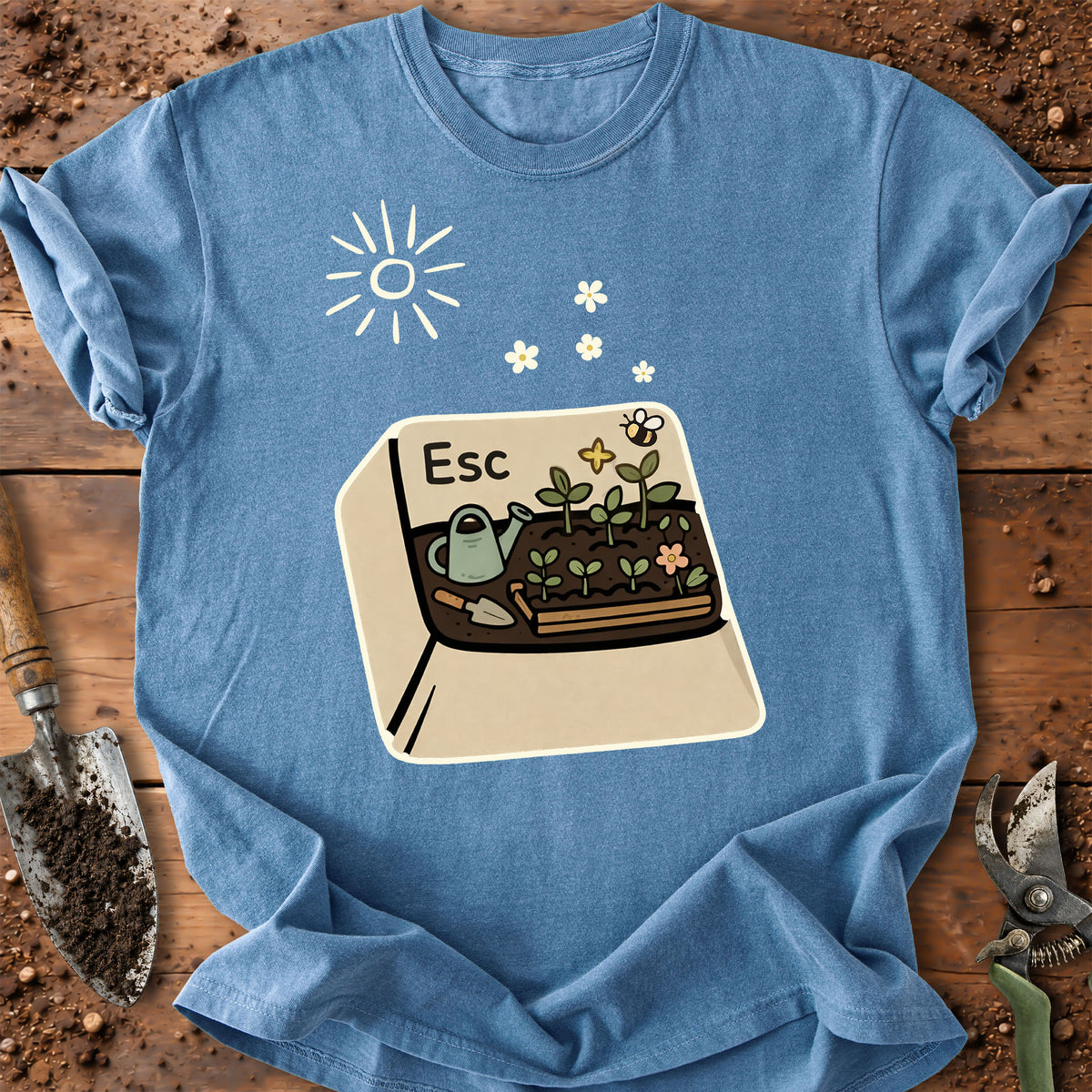 A light blue graphic t-shirt laid flat on a rustic wooden surface with gardening props like a trowel and pruning shears. The shirt features a cartoon graphic of a garden bed with a watering can, flowers, a bee, and the letter 'Esc' printed on a beige square, with a white sun icon above.