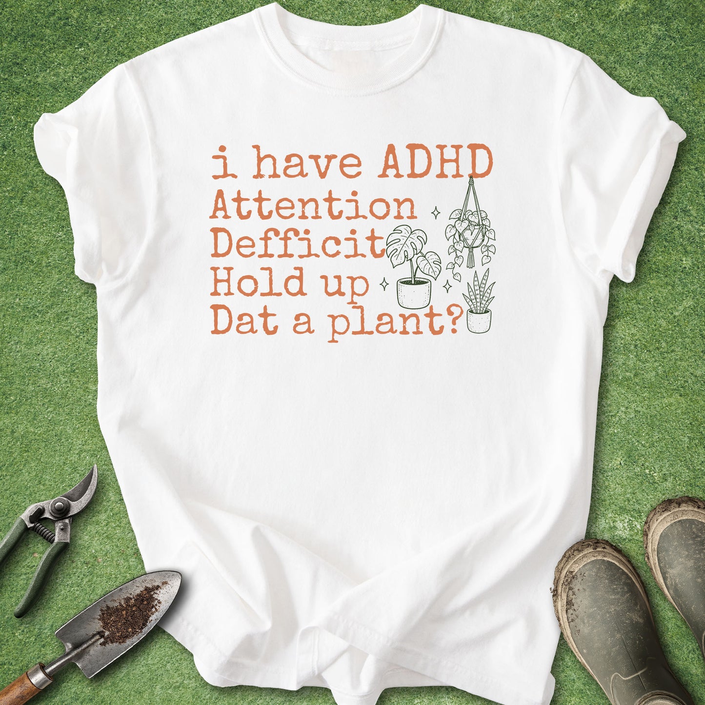 A white short-sleeve graphic T-shirt laid flat on green grass-like background, with orange text reading 'i have ADHD Attention Deficit Hold up Dat a plant?' and small plant illustrations. Gardening tools and muddy boots are partially visible around the shirt.