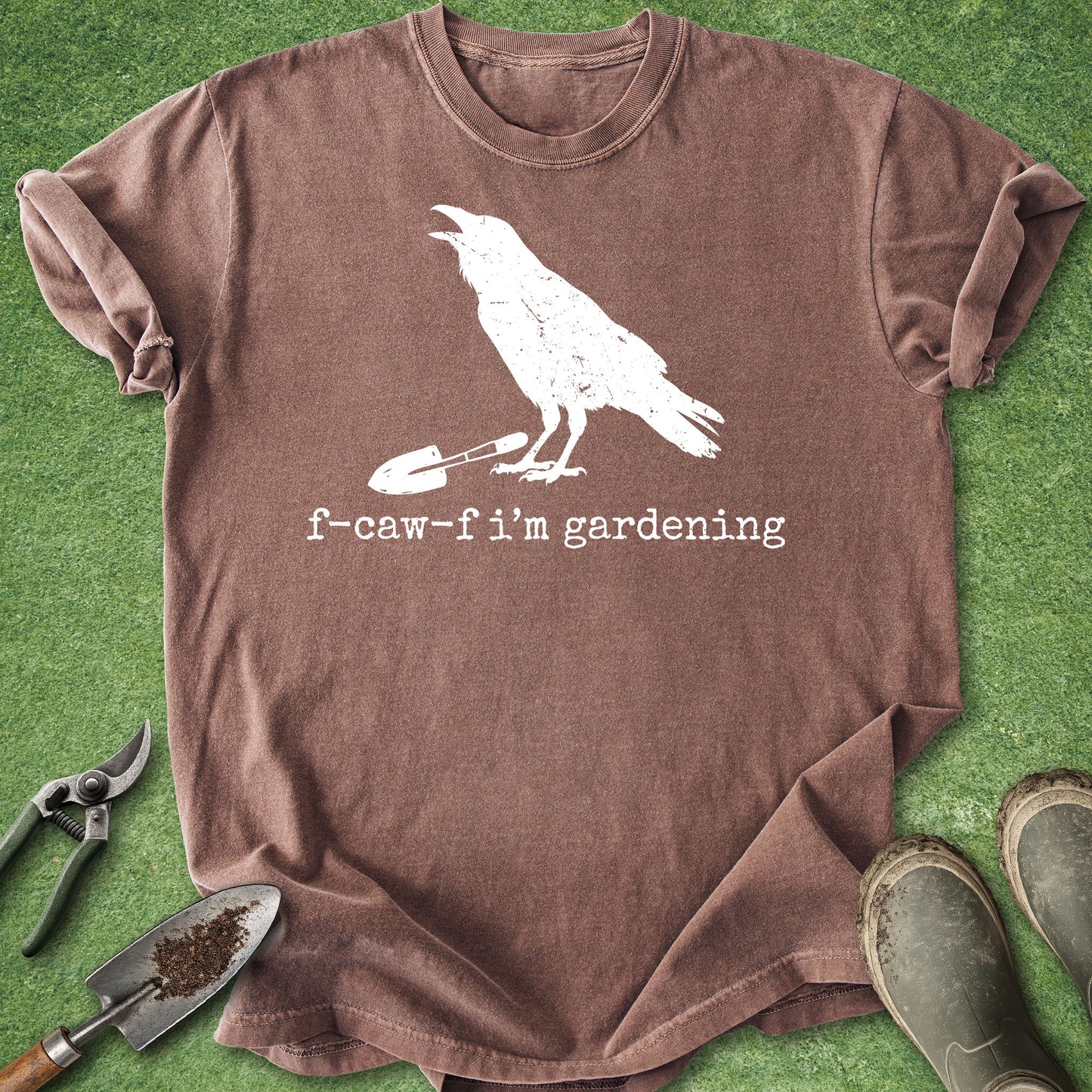 A brown short-sleeve t-shirt with a white graphic of a raven standing on a gardening tool, with the text 'f-caw-f i'm gardening' underneath. The shirt is laid flat on green grass with gardening props including pruning shears, a trowel, and boots.