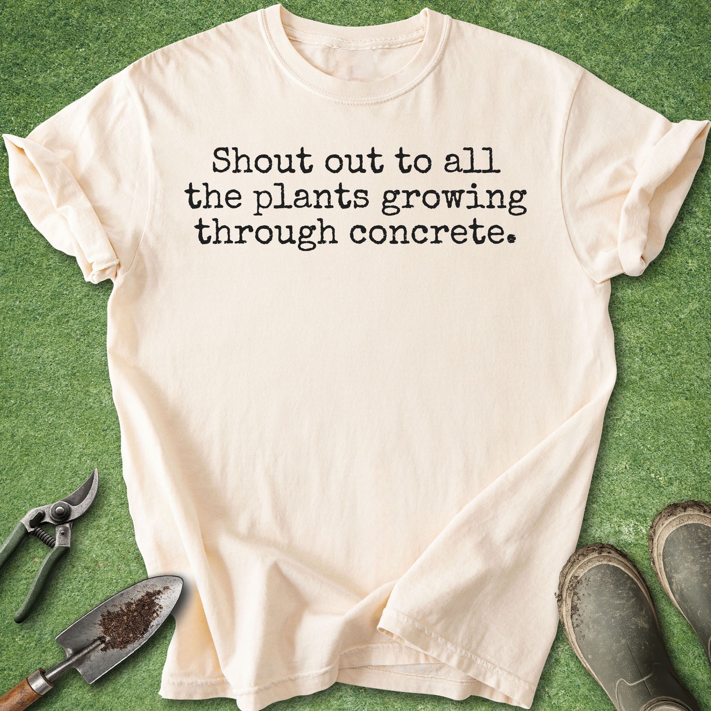 A cream-colored short-sleeve t-shirt with black printed text reading 'Shout out to all the plants growing through concrete.' laid flat on a green grass-like surface, styled with gardening tools and muddy boots around it.