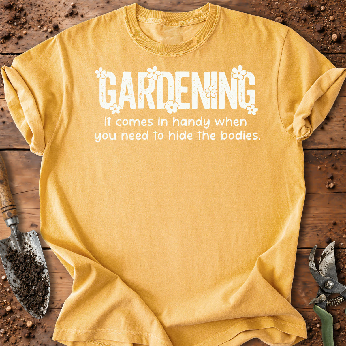 A yellow short-sleeve t-shirt with a white graphic text that reads 'GARDENING It comes in handy when you need to hide the bodies.' styled in a flat-lay on a rustic wooden background with gardening tools.