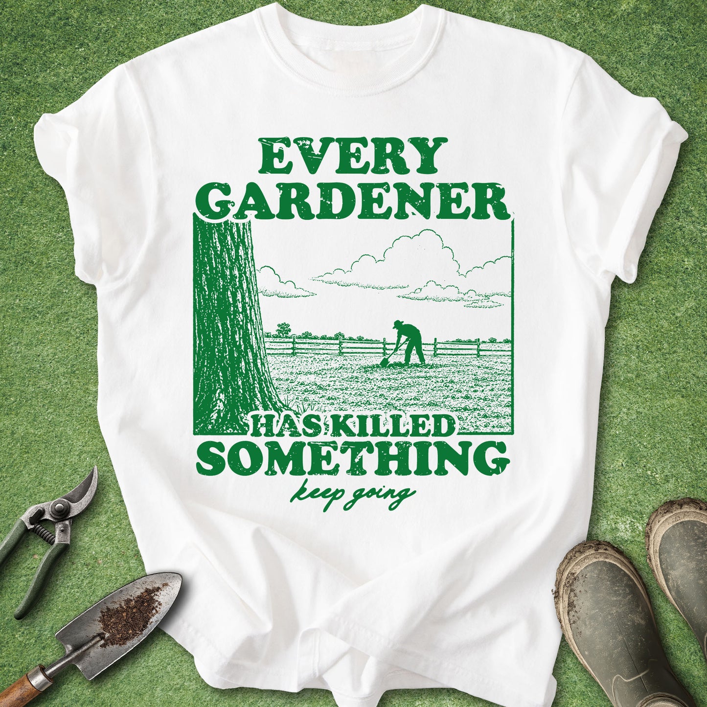 A white short-sleeve graphic T-shirt laid flat on a green grass-like surface, with gardening tools and boots around it. The shirt has a large green printed design and text reading “EVERY GARDENER HAS KILLED SOMETHING keep going.