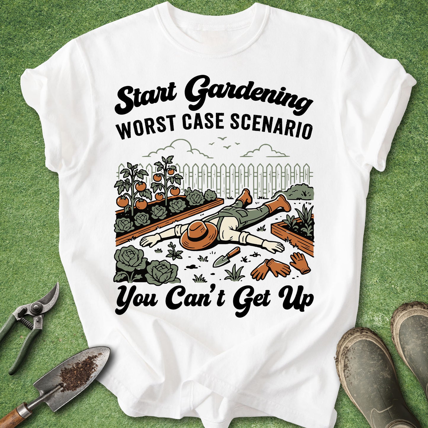 A white graphic t-shirt laid flat on a green grass-like surface, surrounded by gardening props including pruning shears, a trowel, and gardening boots. The shirt features a humorous illustration of a gardener lying on their back in a garden bed with the text 'Start Gardening WORST CASE SCENARIO You Can't Get Up'.