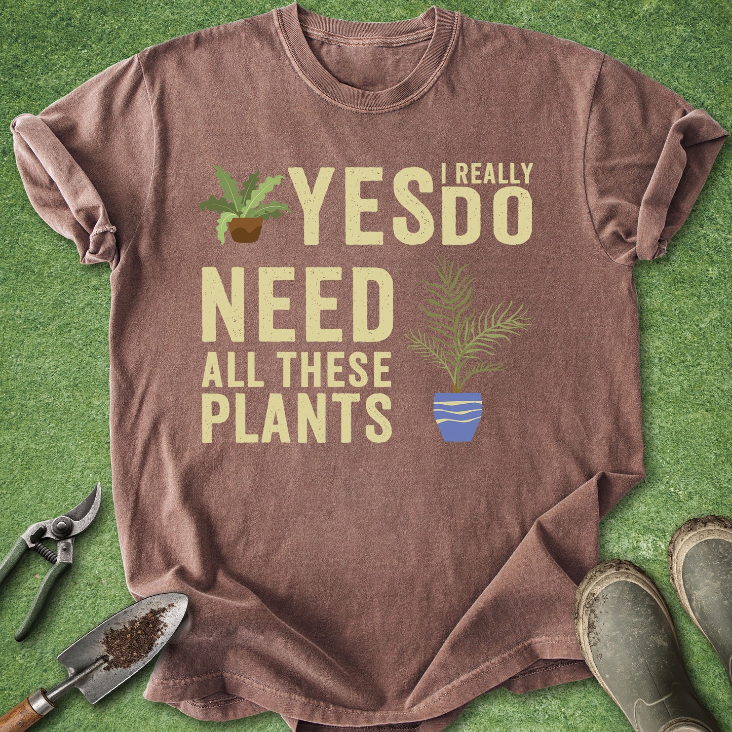 Brown t-shirt with 'I really need all these plants' text and plant graphics on a green grass background.