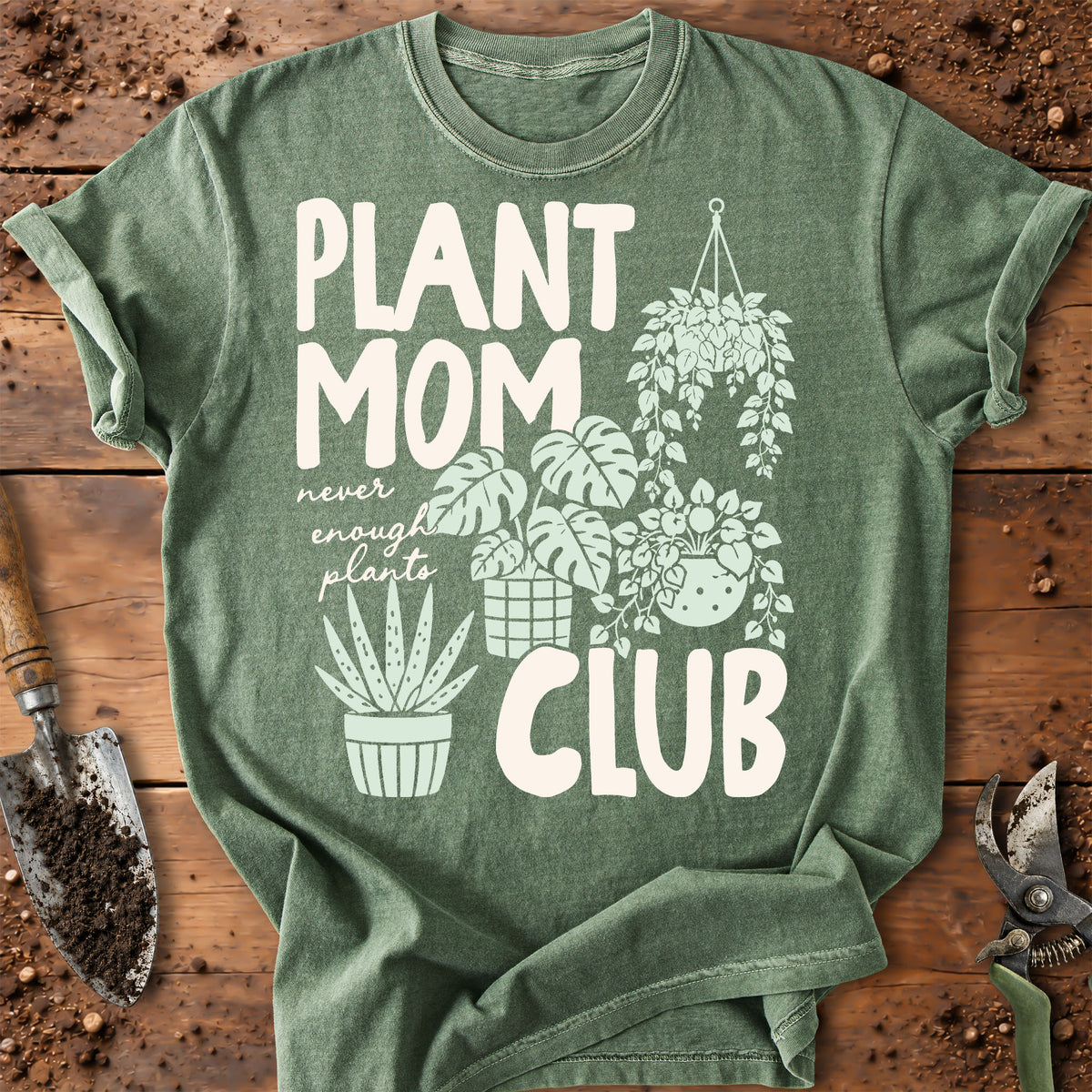 A heather sage green graphic t-shirt laid flat on a rustic wooden surface with gardening props. The shirt features a white graphic print that reads 'PLANT MOM CLUB' with illustrations of various potted and hanging plants.