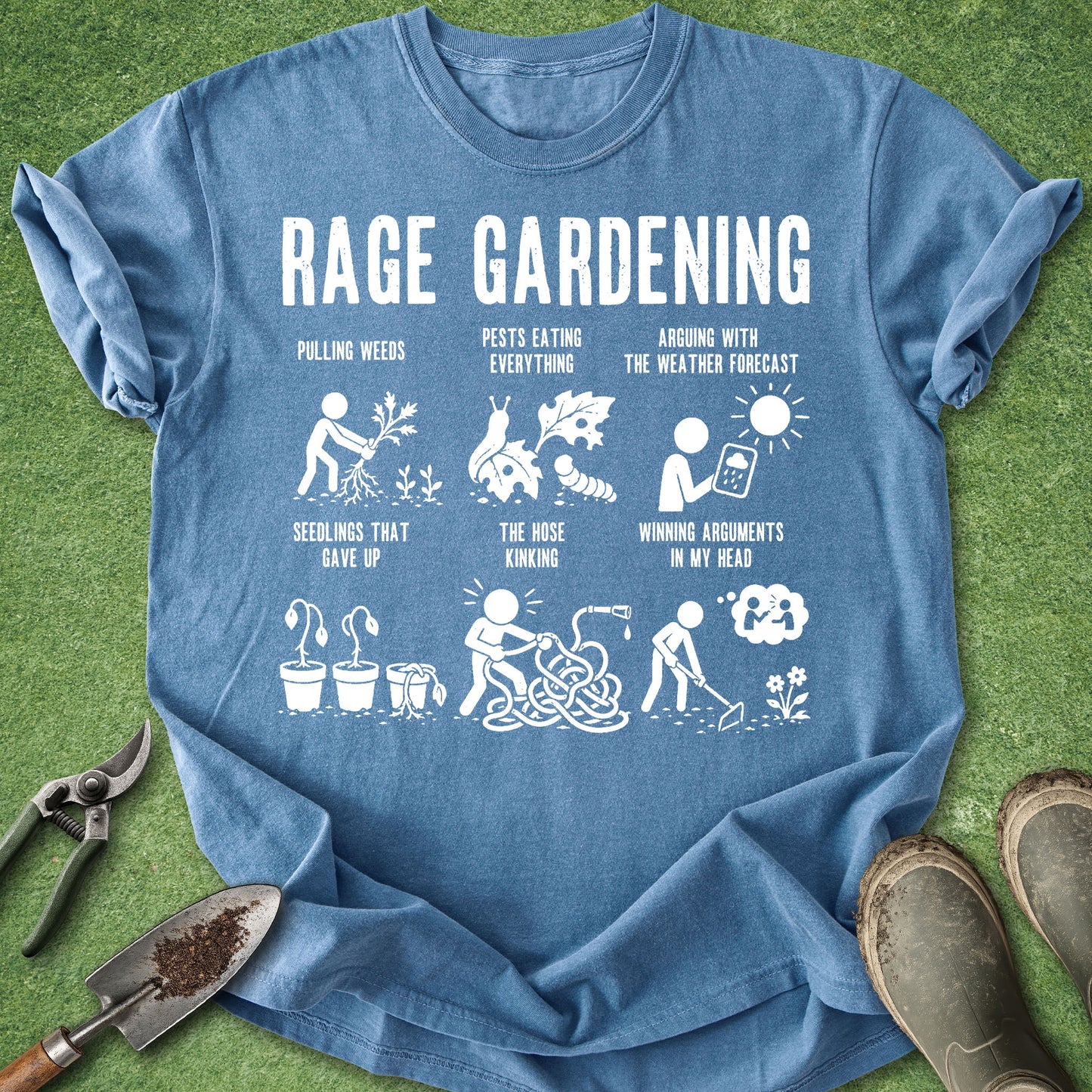 A blue graphic t-shirt laid flat on green grass with gardening tools and boots around it. The shirt features a white 'RAGE GARDENING' print with humorous illustrations and text related to gardening.