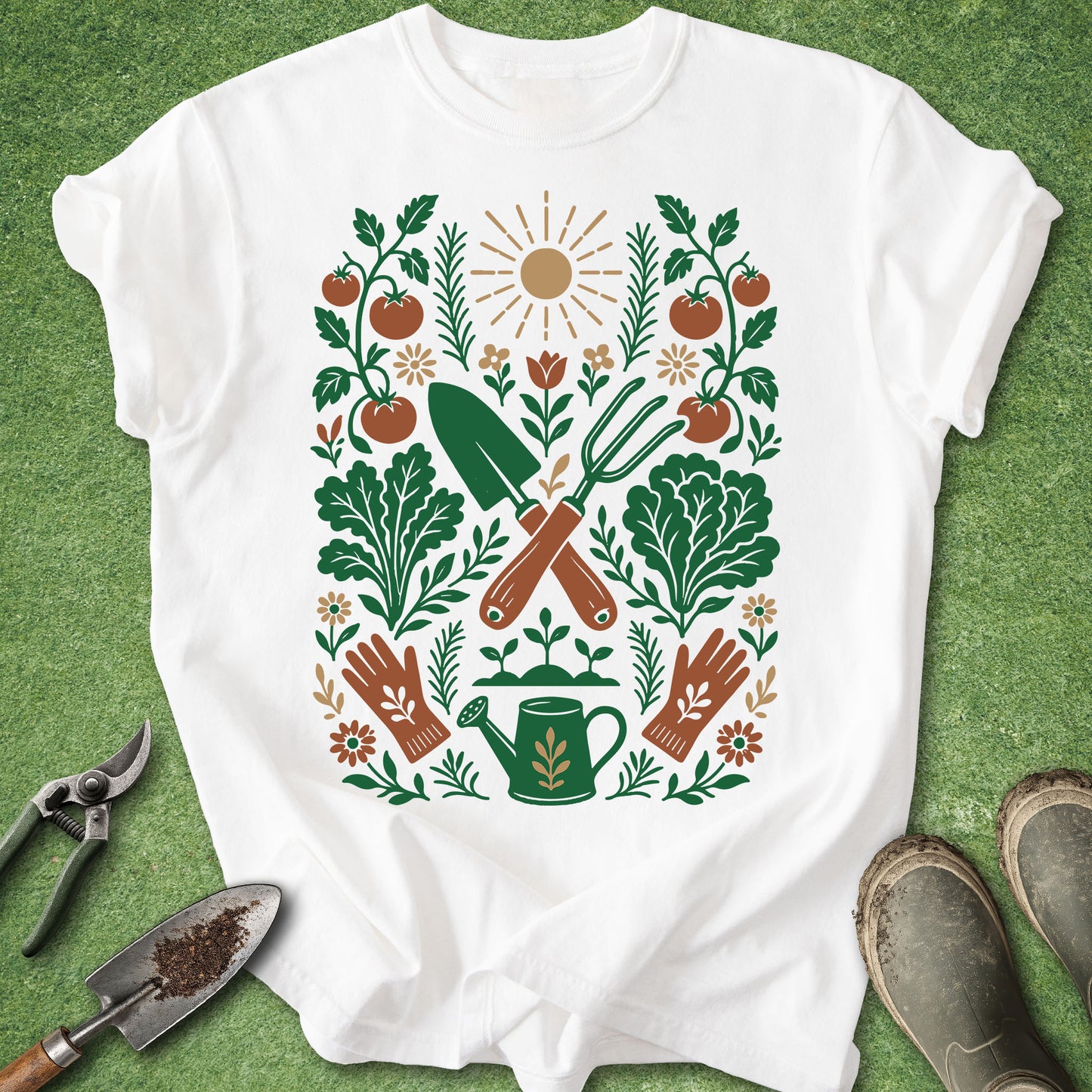A white short-sleeve T-shirt laid flat on a green grass-like surface, featuring a large colorful gardening graphic with tomatoes, leaves, gloves, a watering can, and tools. Gardening props including shears, a trowel, and boots are placed around the shirt.