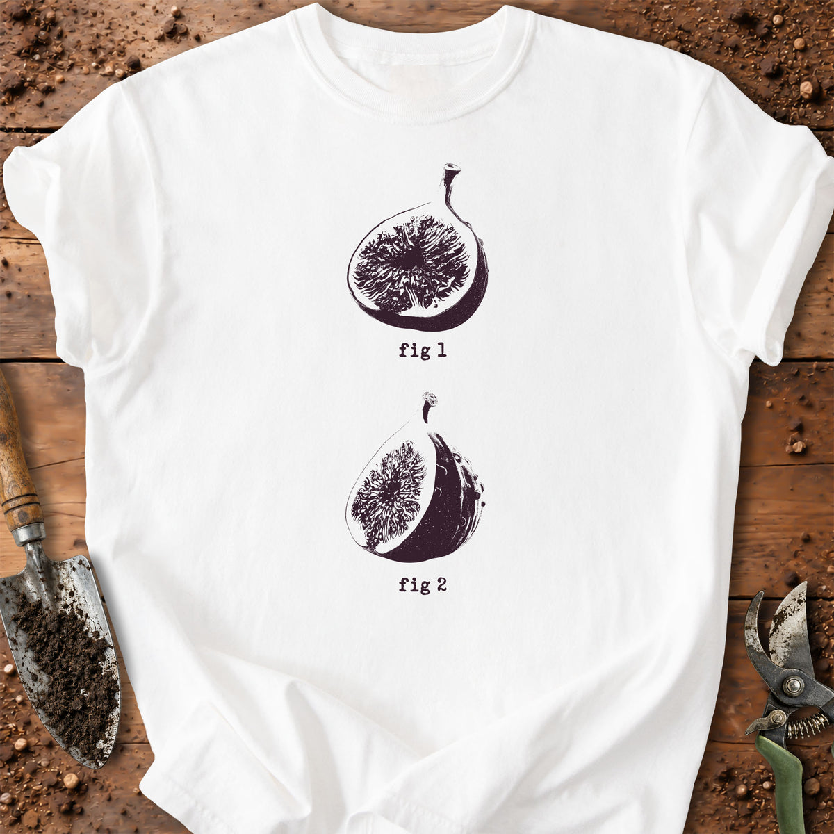 A white short-sleeve T-shirt laid flat on a rustic wooden surface with gardening props. The shirt has two black illustrated fig halves with the words 'fig 1' and 'fig 2' printed beneath each.