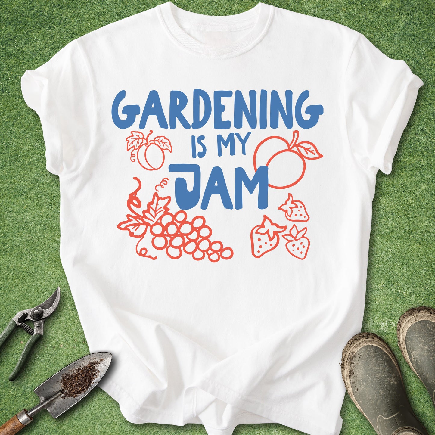 A white short-sleeve graphic T-shirt laid flat on green grass, with the phrase 'GARDENING IS MY JAM' printed in blue and red fruit illustrations. Gardening tools and boots are partially visible around the shirt.