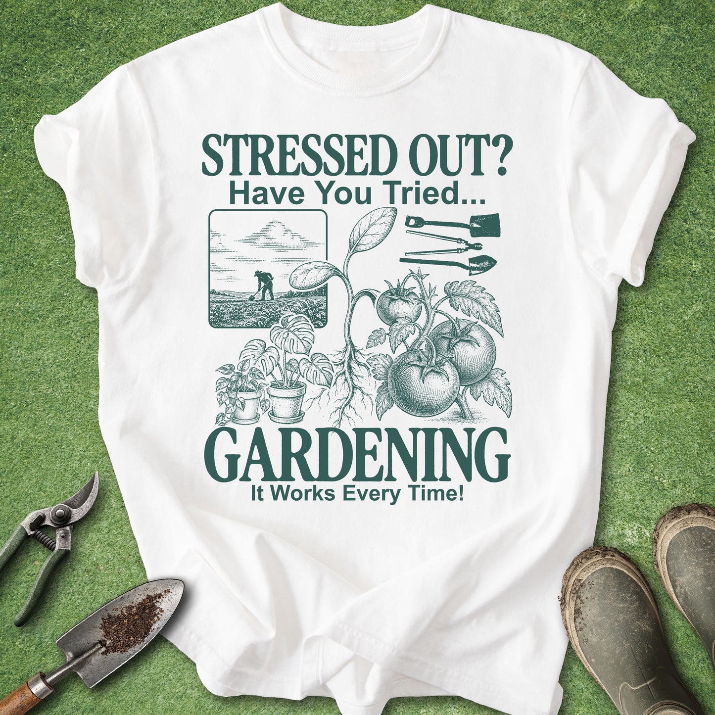 A white short-sleeve graphic T-shirt laid flat on green grass-like background, with gardening-themed printed text and illustrations including tomatoes, plants, tools, and a small dog scene. Gardening props such as pruning shears, a trowel, and boots are placed around the shirt.