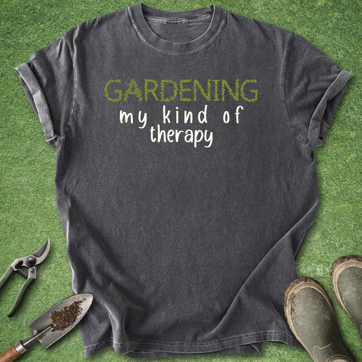 A dark charcoal gray t-shirt with the text 'GARDENING my kind of therapy' printed on the chest, styled in a flat-lay on green grass with gardening tools (trowel, pruning shears) and boots.