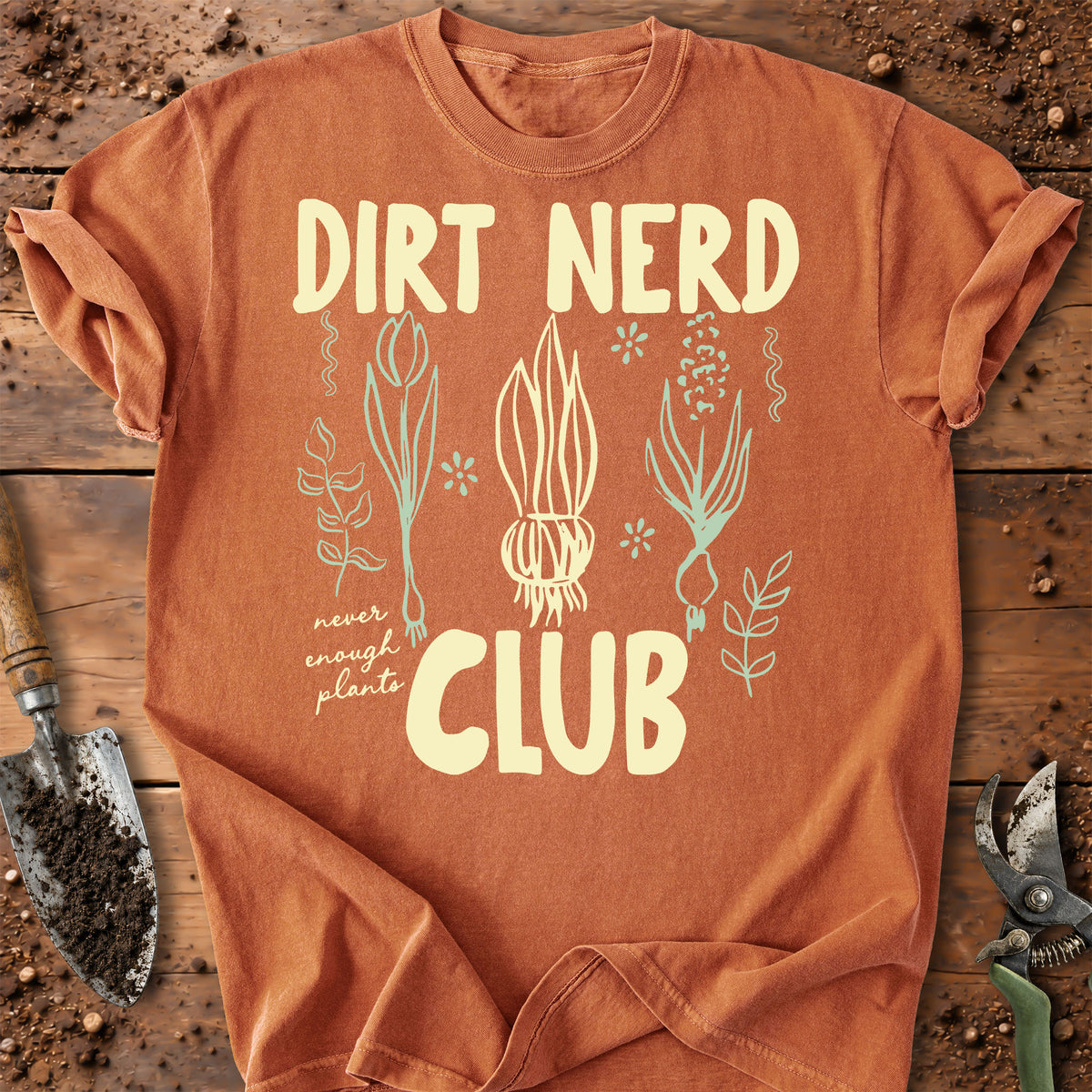 An orange short-sleeve t-shirt with a 'Dirt Nerd Club' graphic, laid flat on a rustic wooden background with gardening tools like a trowel and pruning shears.