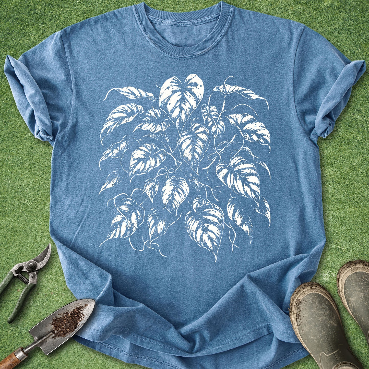 A blue short-sleeve crewneck t-shirt with a white leaf graphic print, laid flat on green grass with gardening tools and boots nearby.