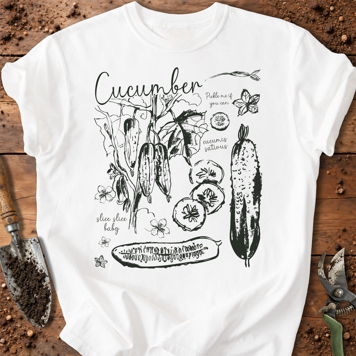 A white short-sleeve T-shirt laid flat on a rustic wooden surface with gardening props. The shirt features a black hand-drawn style graphic of cucumbers, leaves, flowers, and text including 'Cucumber' and 'Pickle me if you can.