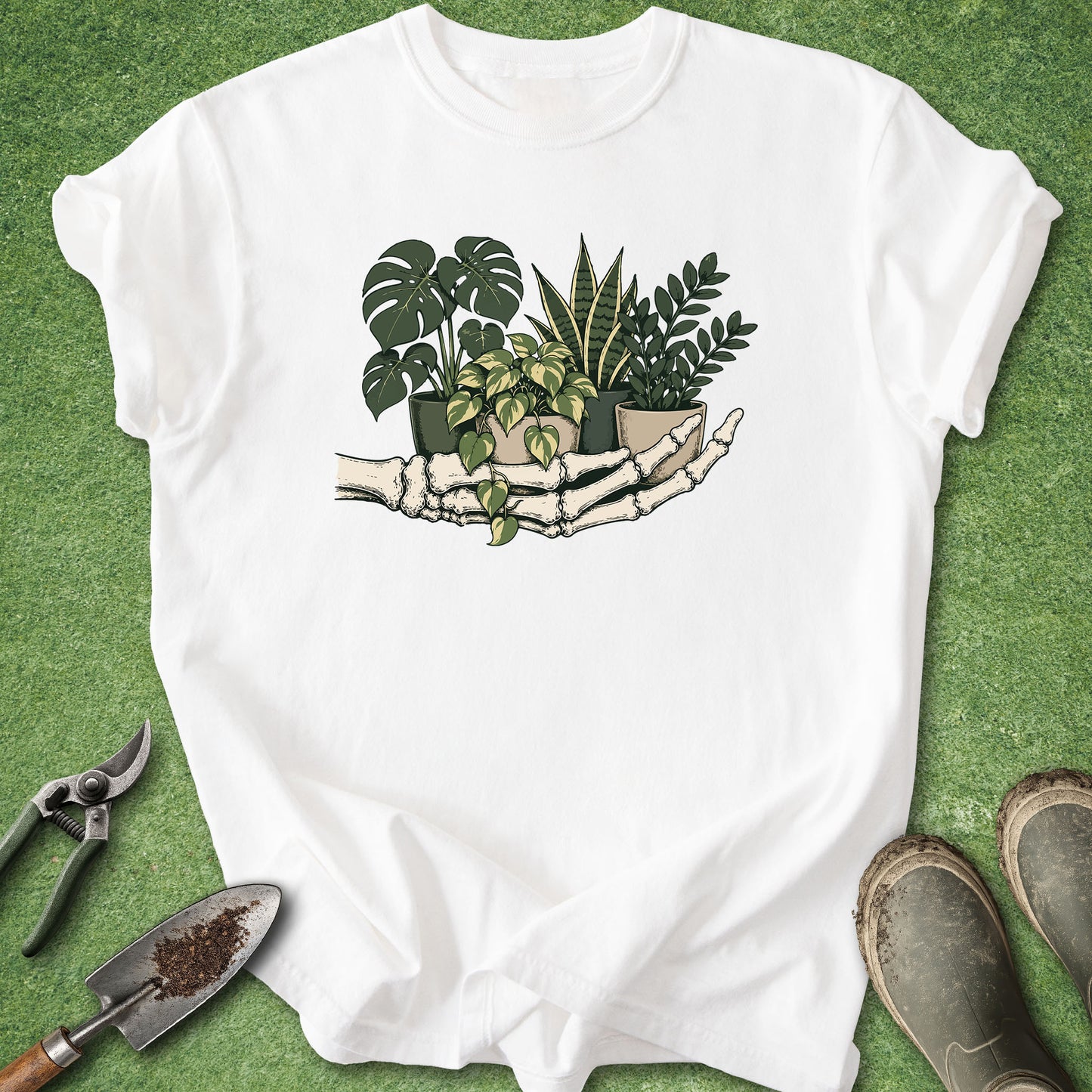 A white short-sleeve T-shirt laid flat on green grass-like background, featuring a central graphic of a skeleton hand holding several potted green plants. Gardening props including pruning shears, a trowel, and boots are placed around the shirt.