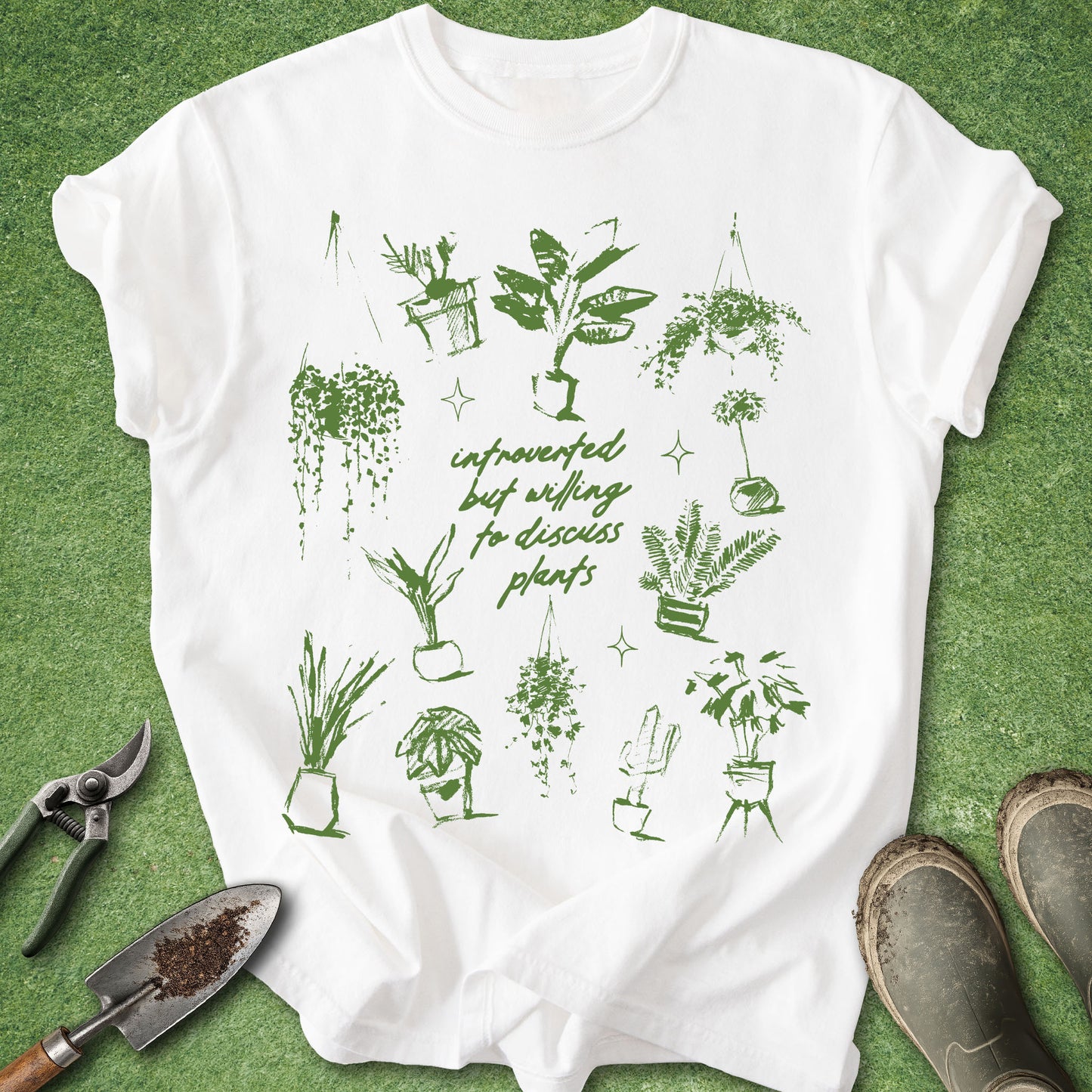 A white short-sleeve graphic T-shirt laid flat on green grass-like artificial turf, surrounded by gardening props including pruning shears, a small hand trowel, and muddy boots. The shirt has a green illustrated print of multiple potted houseplants and cursive text reading 'infraordinary but willing to discuss plants.