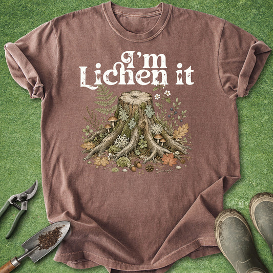 A brown short-sleeve t-shirt laid flat on green grass, featuring a white graphic that reads 'I'm Lichen it' above an illustration of a tree stump with mushrooms and greenery. Gardening tools and boots are placed around the shirt.