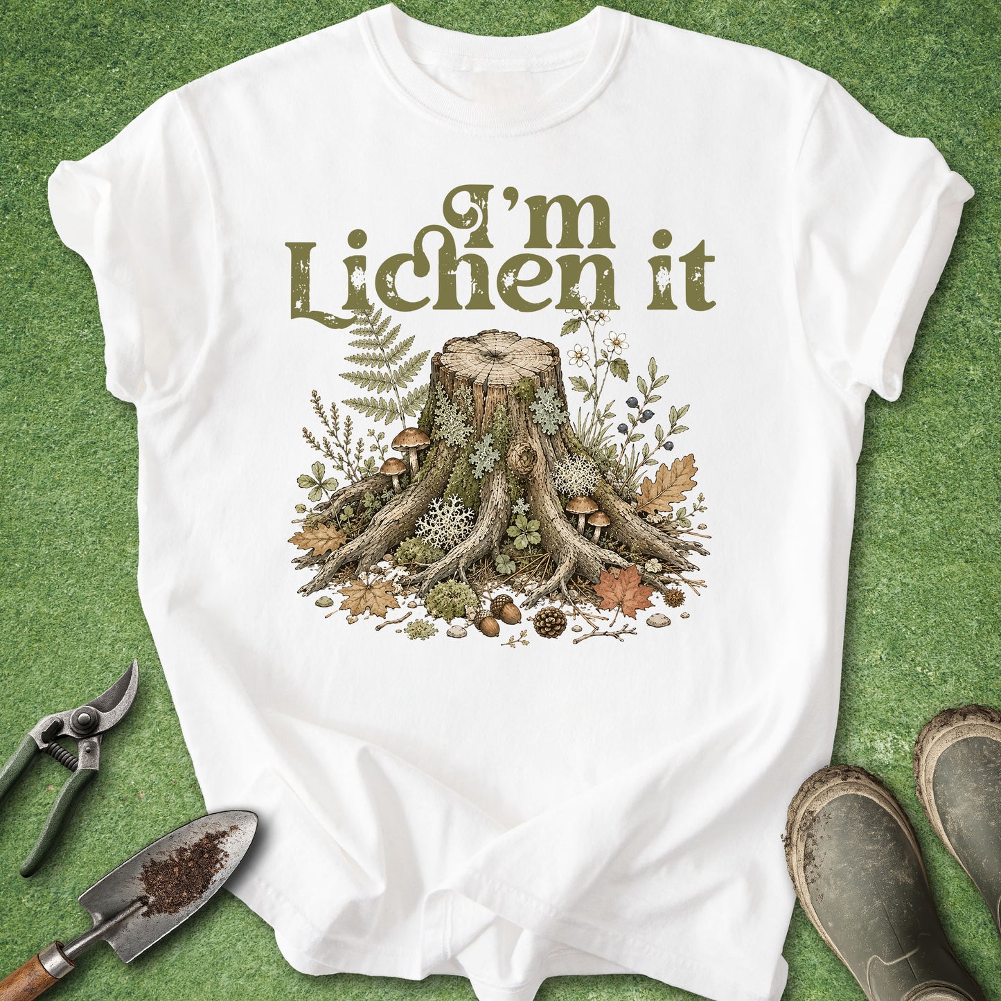A white short-sleeve graphic T-shirt laid flat on green grass, featuring the phrase “I’m Lichen it” above an illustrated tree stump with roots, moss, ferns, and small mushrooms. Gardening props including pruning shears, a trowel, and boots are placed around the shirt.