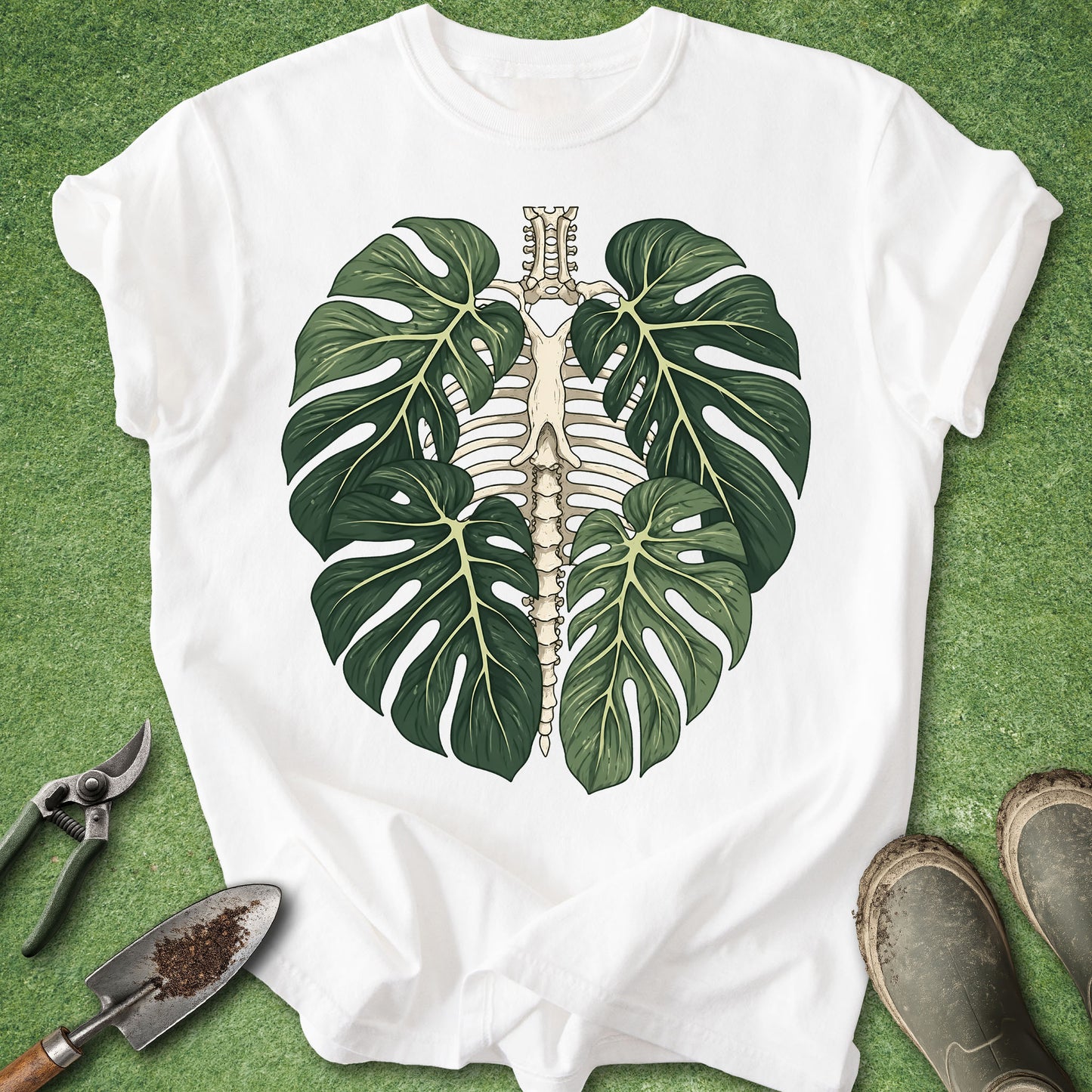 A white short-sleeve T-shirt laid flat on green grass, featuring a large graphic of two green monstera leaves framing a beige rib cage and spine illustration. Gardening tools and muddy boots are placed around the shirt.