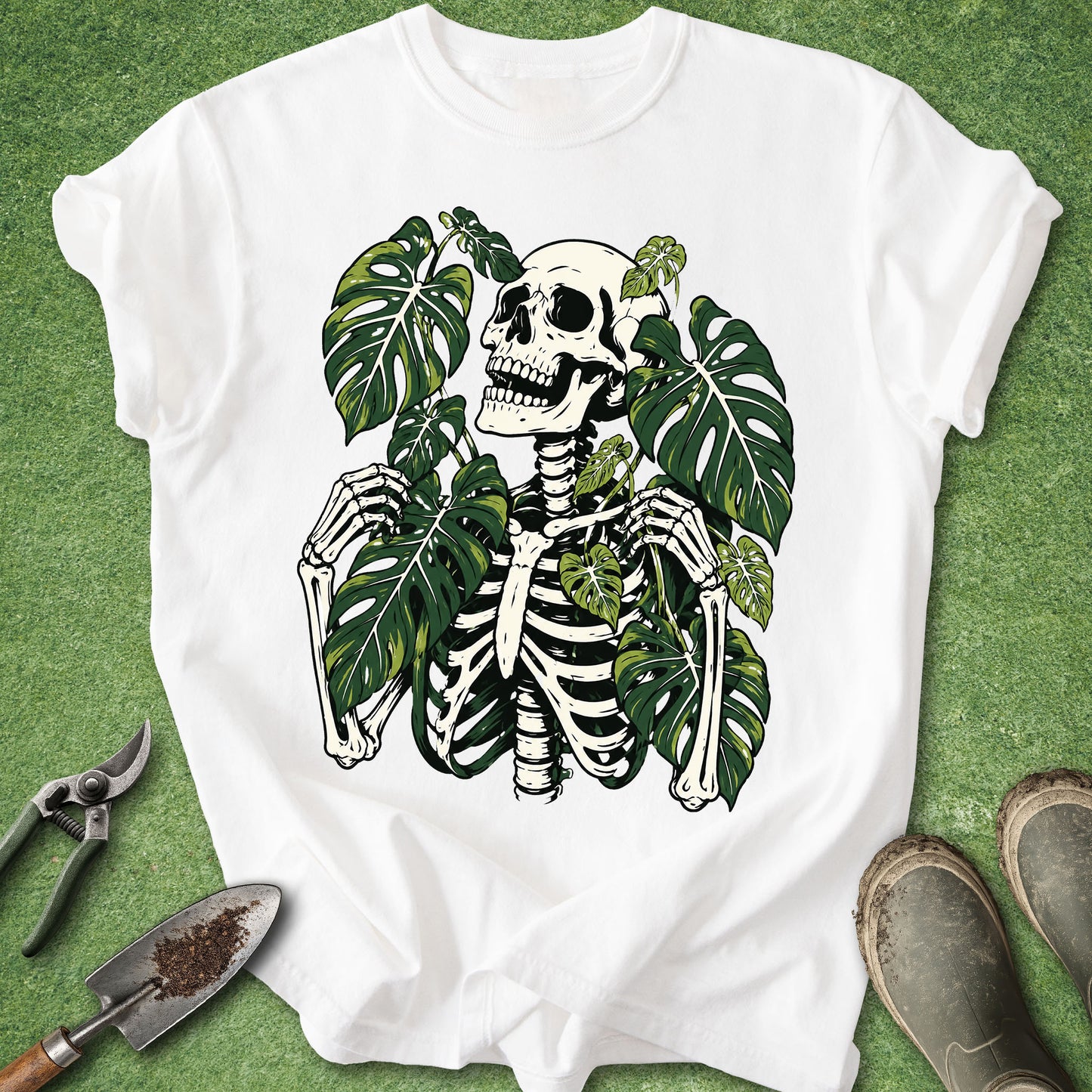 A white short-sleeve T-shirt laid flat on green grass, featuring a large graphic of a black-and-white skeleton surrounded by green monstera leaves. Gardening tools and boots are partially visible around the shirt as styling props.
