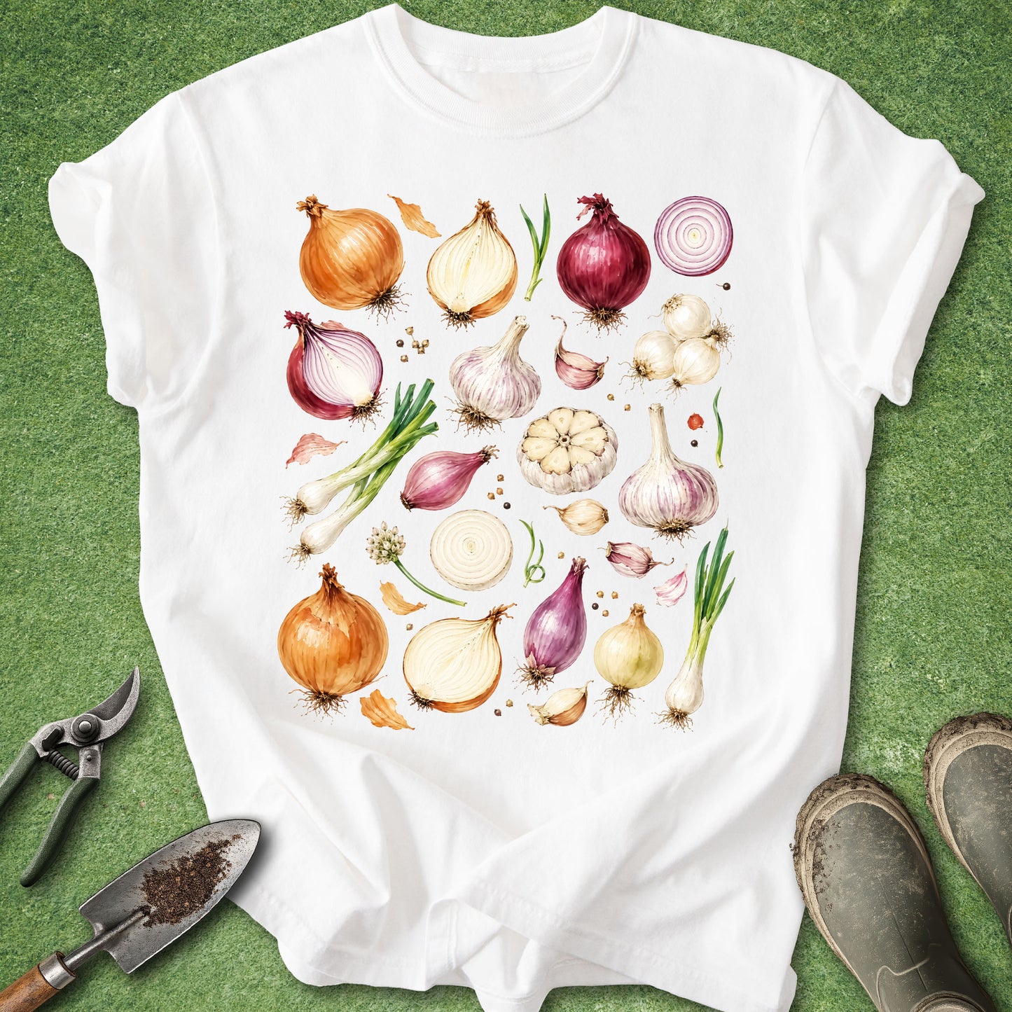 A white short-sleeve crewneck T-shirt laid flat on green grass, featuring a colorful watercolor-style print of various onions, garlic bulbs, scallions, and peppercorns on the front. Gardening props, including a trowel, pruning shears, and boots, are visible around the shirt.
