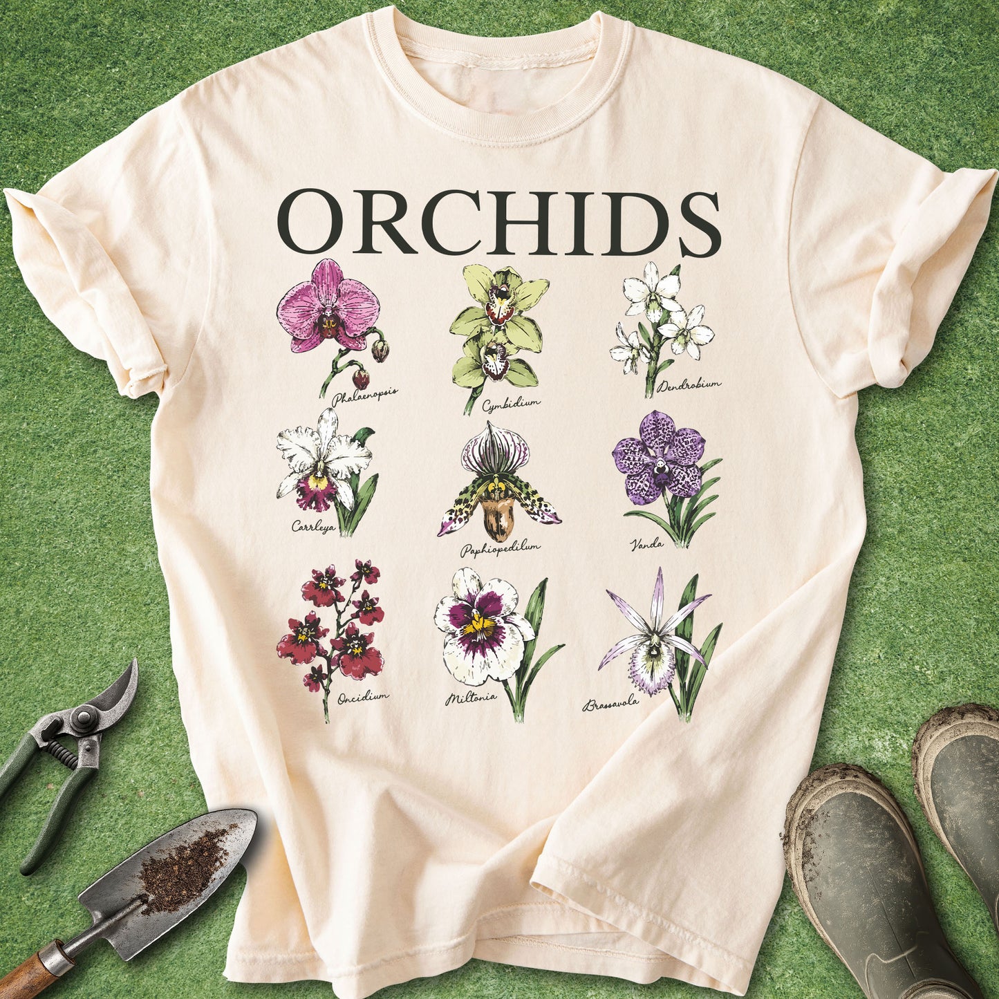 A cream-colored short-sleeve t-shirt with a graphic print of various orchid flowers and the word 'ORCHIDS' in bold black letters. The shirt is laid flat on a green grass-like surface, surrounded by gardening props including a trowel, pruning shears, and boots.