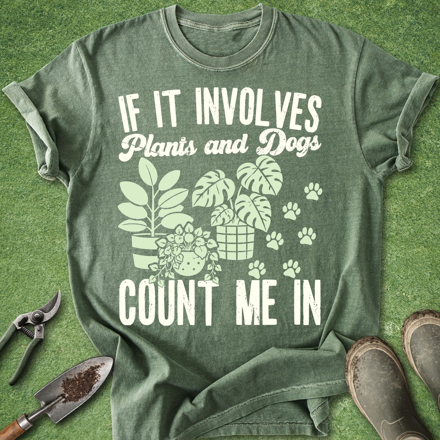 An olive green graphic t-shirt laid flat on artificial grass, featuring the text 'IF IT INVOLVES Plants and Dogs COUNT ME IN' in white, alongside illustrations of potted plants, a dog, and paw prints. Gardening tools and boots are styled around the shirt.