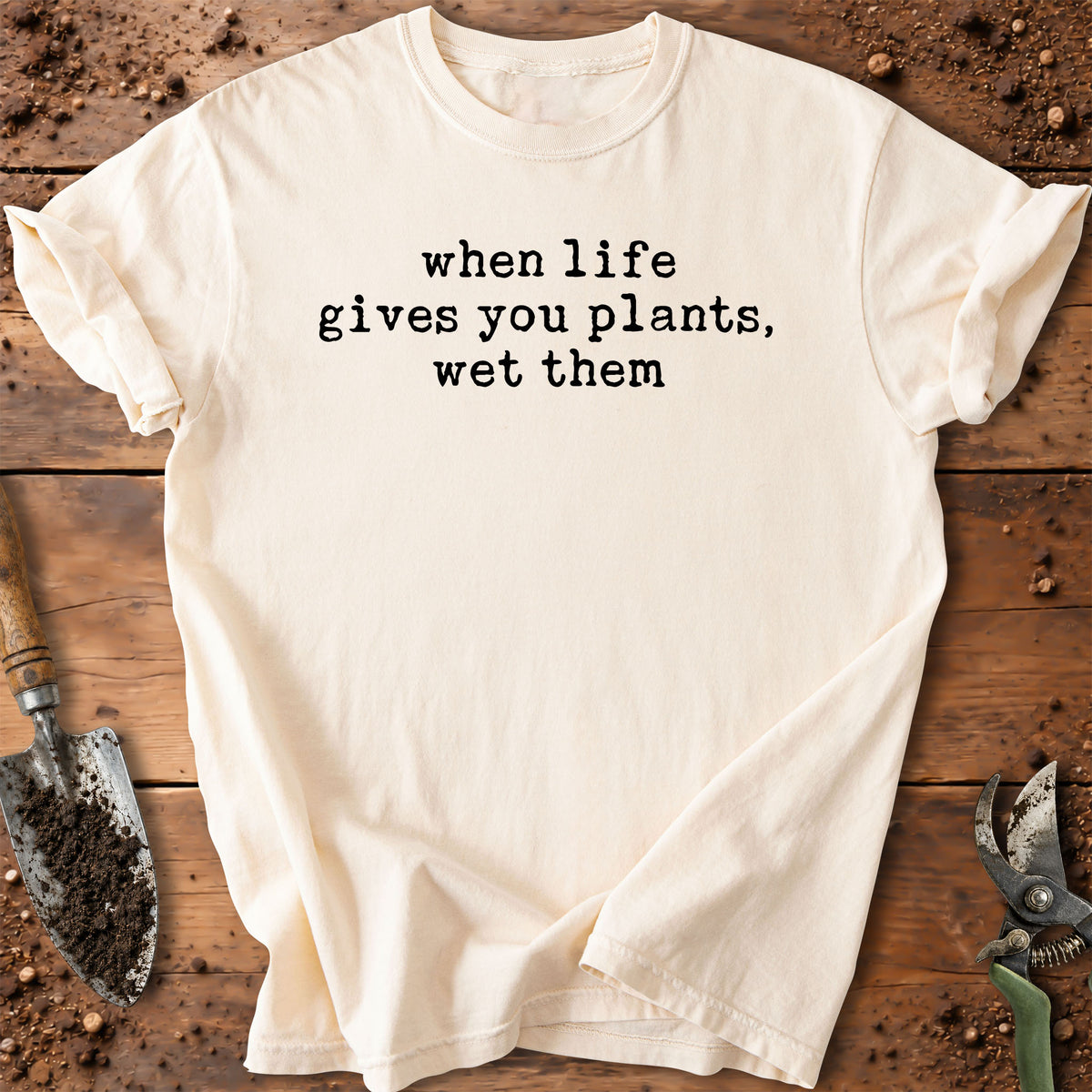 An off-white short-sleeve t-shirt with the black printed text 'when life gives you plants, wet them' laid flat on a rustic wooden surface with gardening tools.