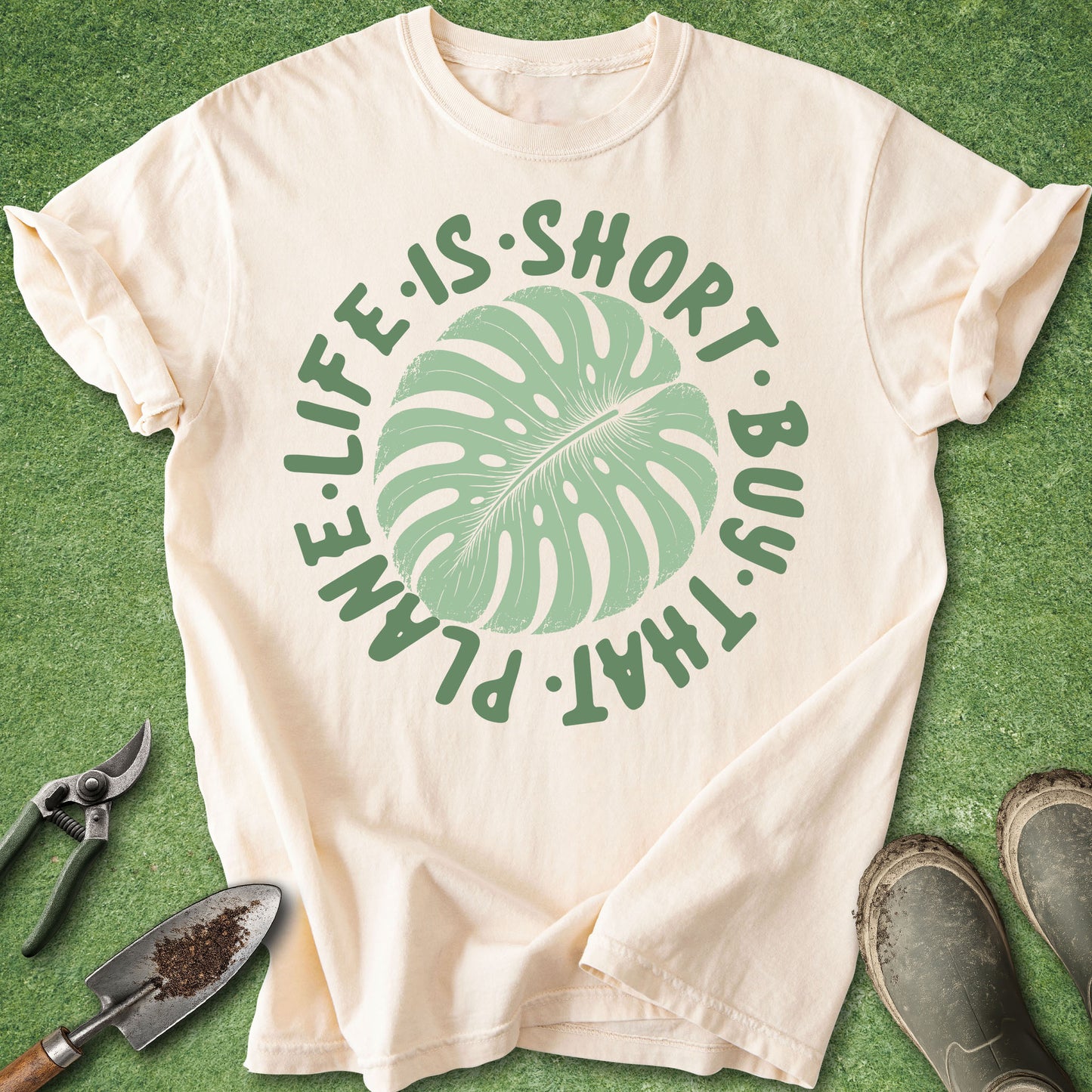 Beige t-shirt with green leaf graphic and text on a green surface with gardening tools.