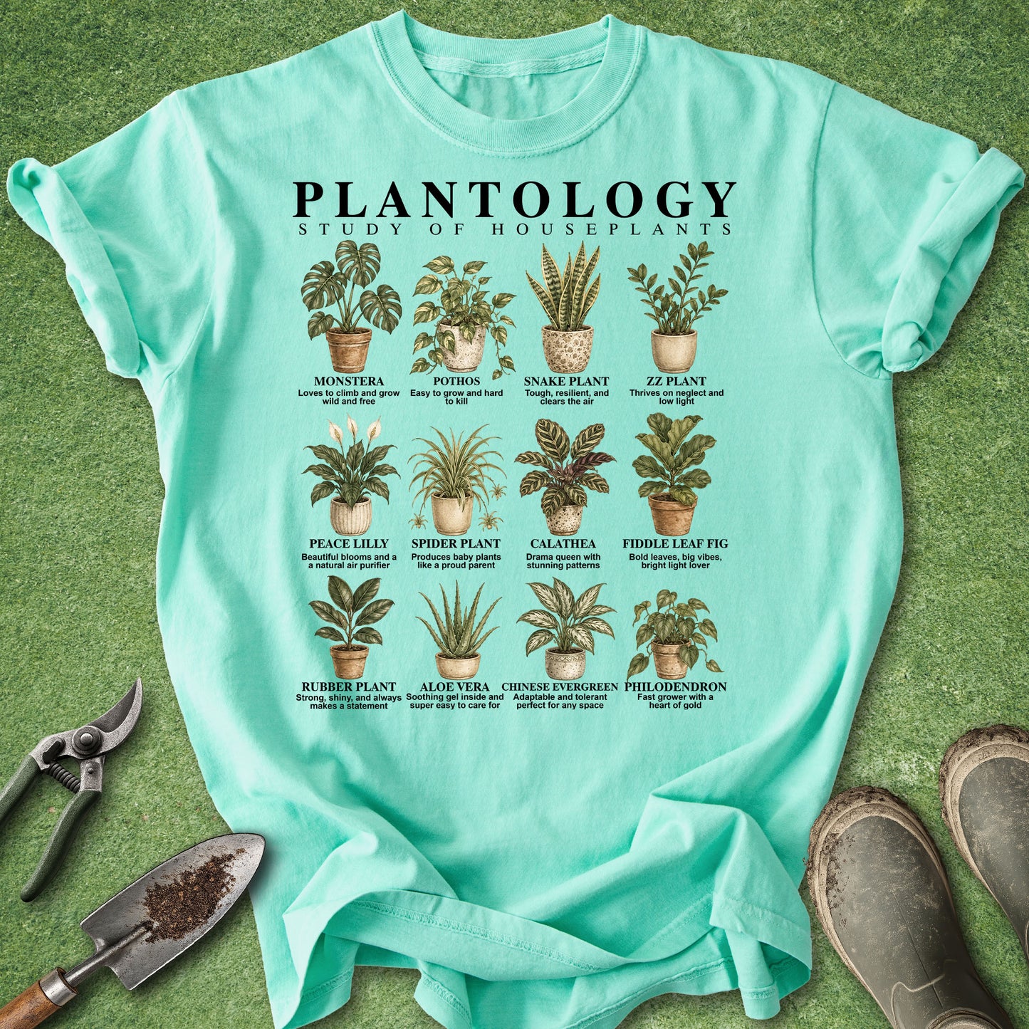 A mint green graphic t-shirt laid flat on a green grass-like surface, surrounded by gardening tools including pruning shears, a trowel, and work boots. The shirt features a detailed print titled 'PLANTOLOGY STUDY OF HOUSEPLANTS' with 15 different houseplant illustrations and names.