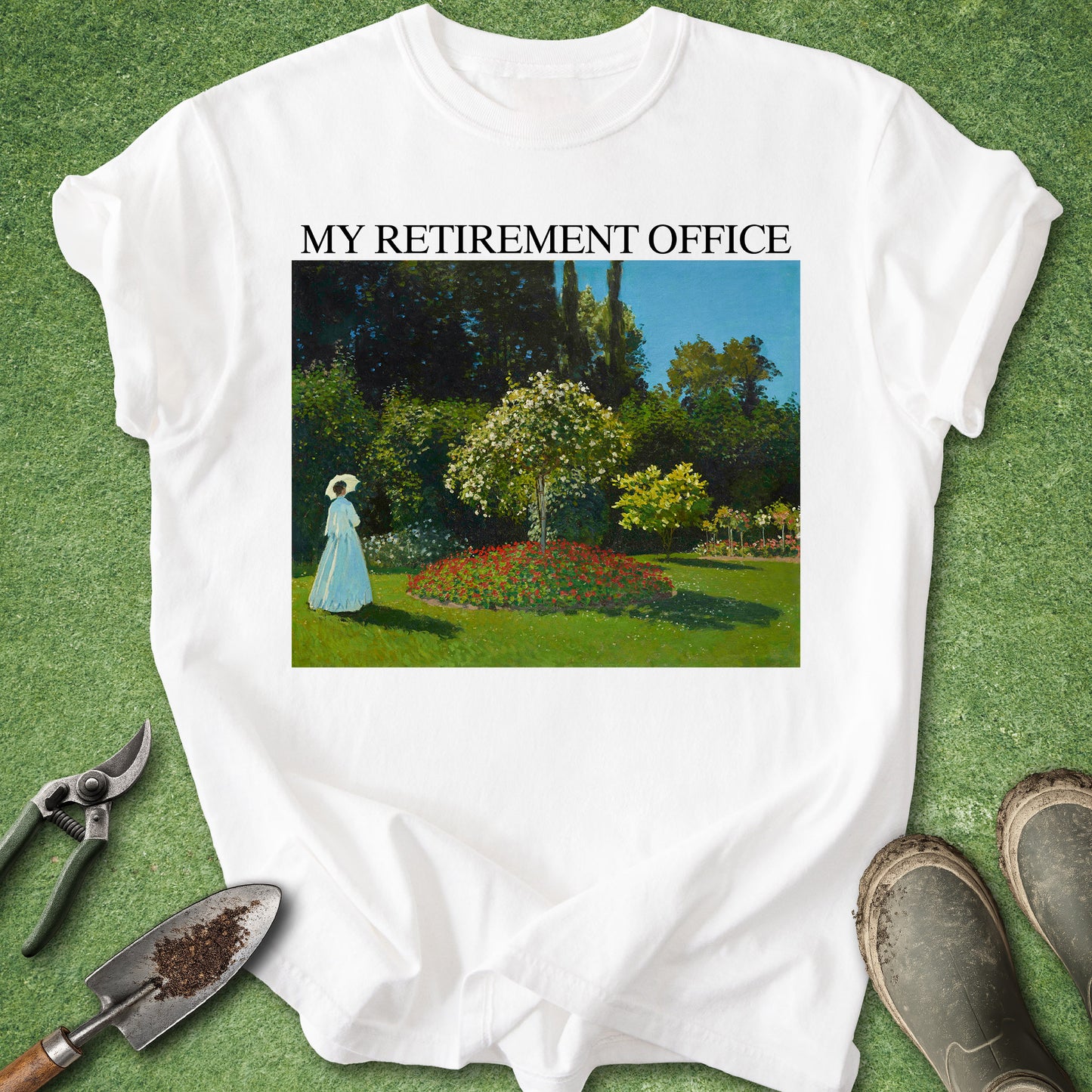 A white short-sleeve graphic T-shirt laid flat on green grass, with the text 'MY RETIREMENT OFFICE' above a painted garden scene. Gardening tools and boots are placed around the shirt as styling props.