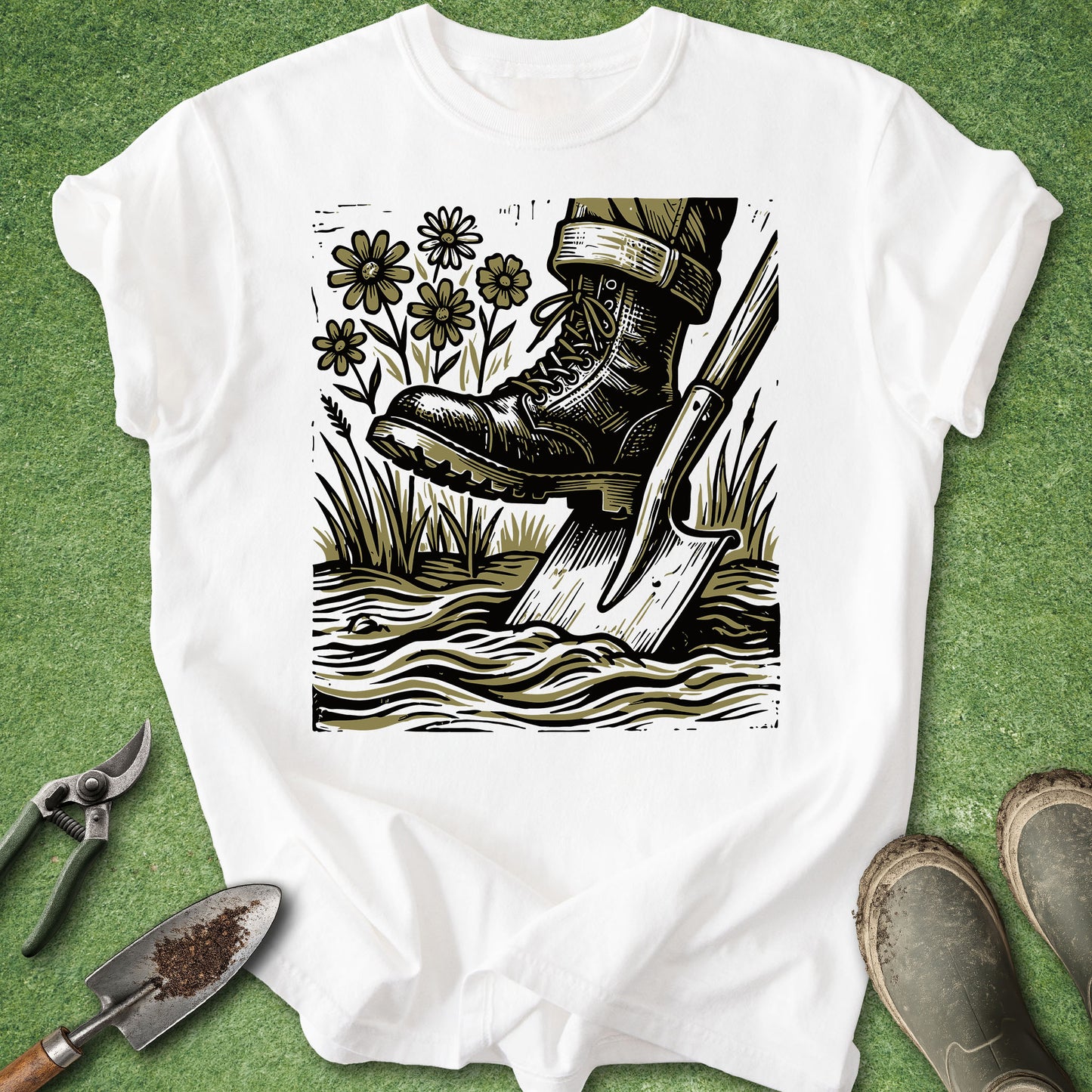 A white short-sleeve T-shirt laid flat on a green textured surface, featuring a large black-and-olive block-print style graphic of a boot stepping into water with a shovel, flowers, and grass. Gardening tools and muddy shoes are placed around the shirt as styling props.