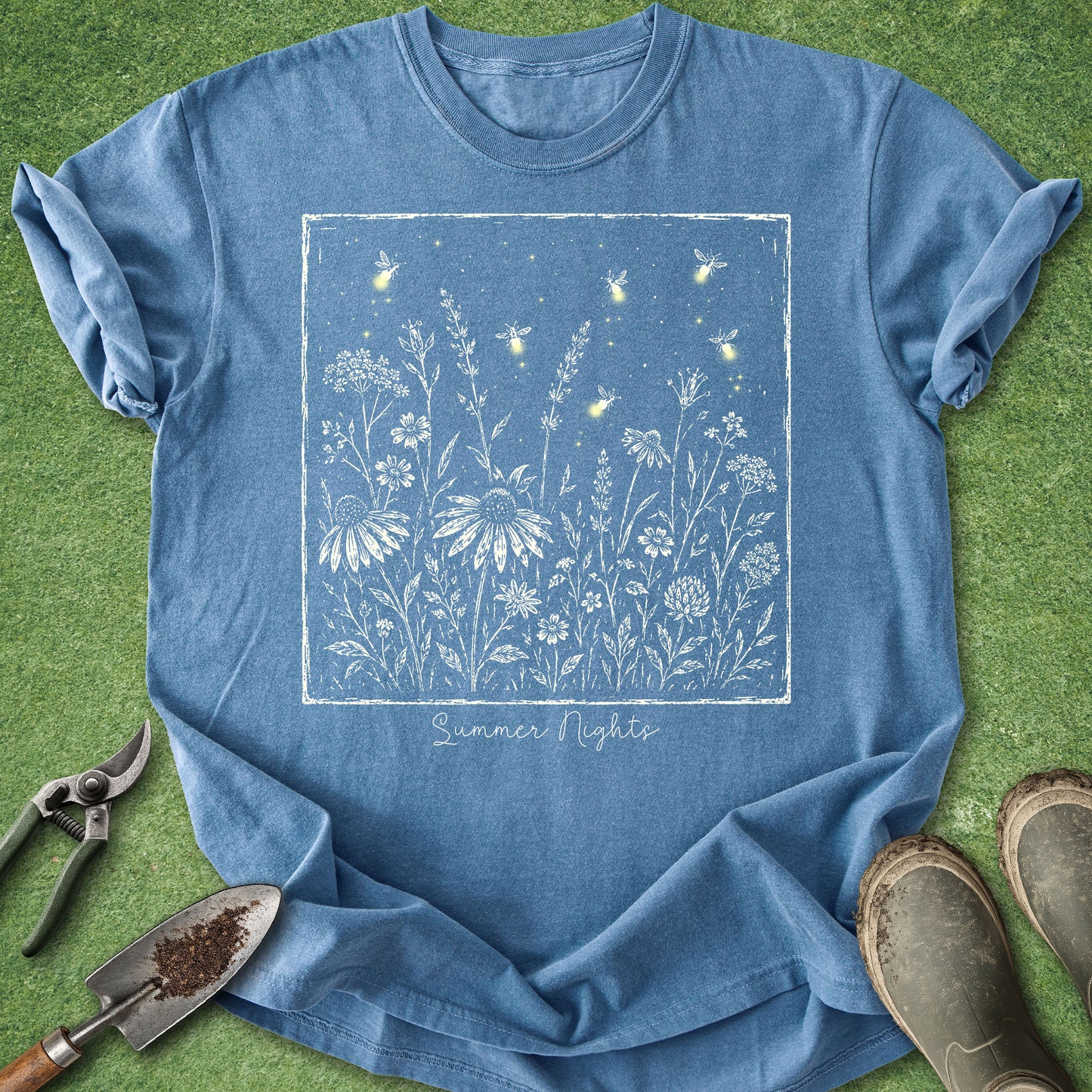 A blue short-sleeve T-shirt laid flat on green grass with gardening tools and boots around it. The shirt has a large square floral and firefly-style graphic in white and light blue on the front, with the words 'Summer Nights' printed below.