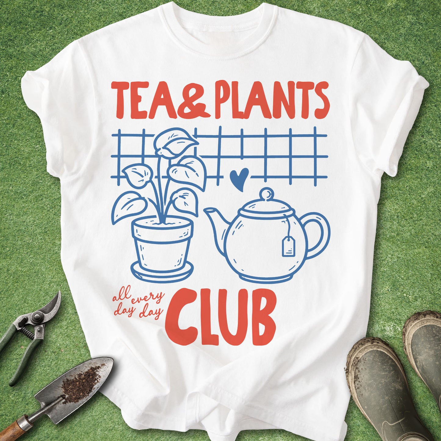 A white graphic T-shirt laid flat on green grass, featuring red and blue text that reads 'TEA & PLANTS all every day CLUB' with a teapot, potted plant, and small heart illustration. Gardening tools and muddy boots are placed around the shirt as styling props.