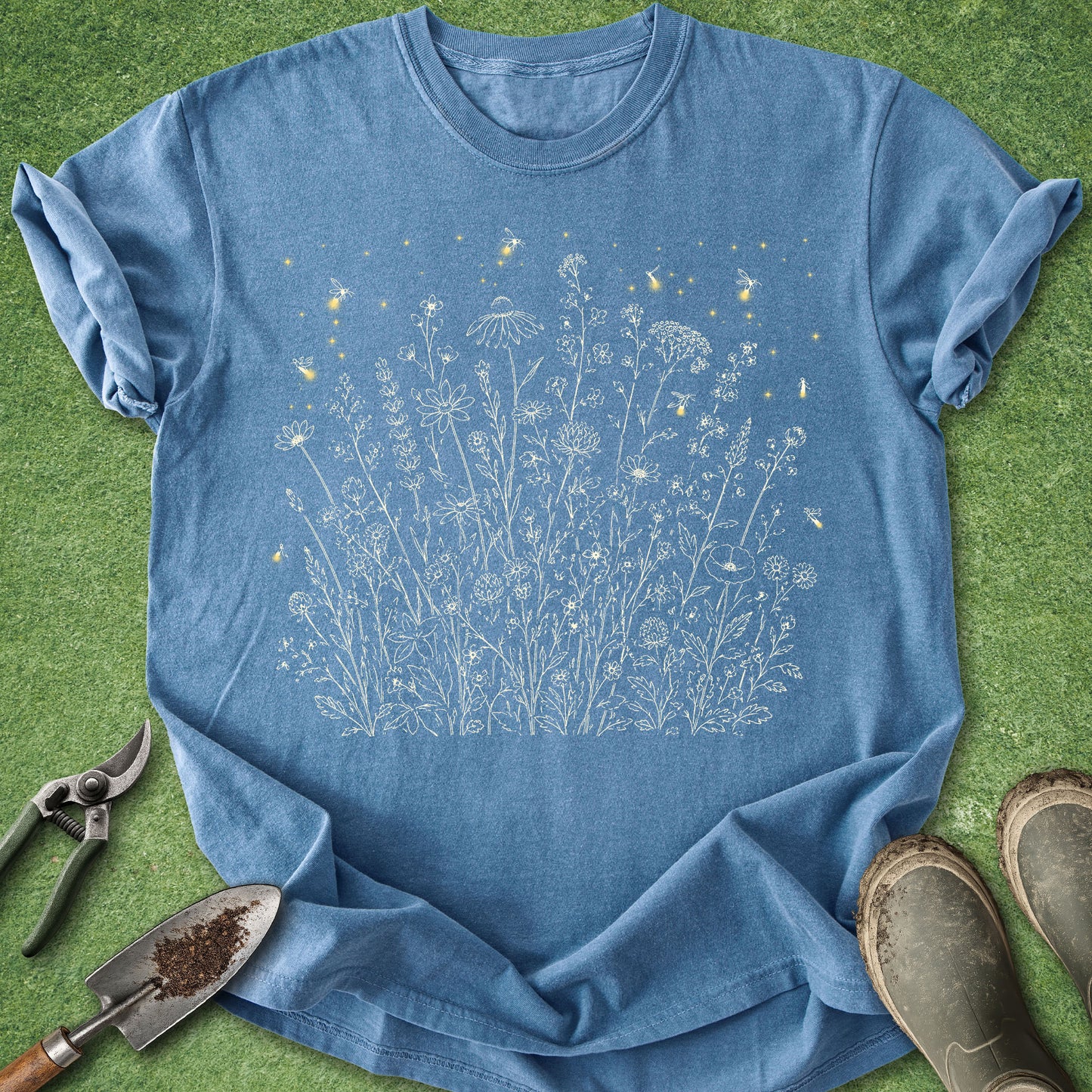 A blue short-sleeve crewneck T-shirt laid flat on green grass, featuring a white line-art graphic of wildflowers and small yellow insects. Gardening tools and muddy boots are placed around the shirt as styling props.
