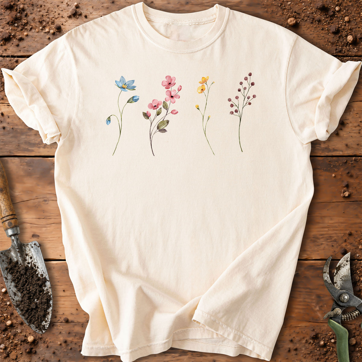An off-white short-sleeve t-shirt with a watercolor-style floral print, laid flat on a rustic wooden surface with gardening tools like a trowel and shears visible around it.