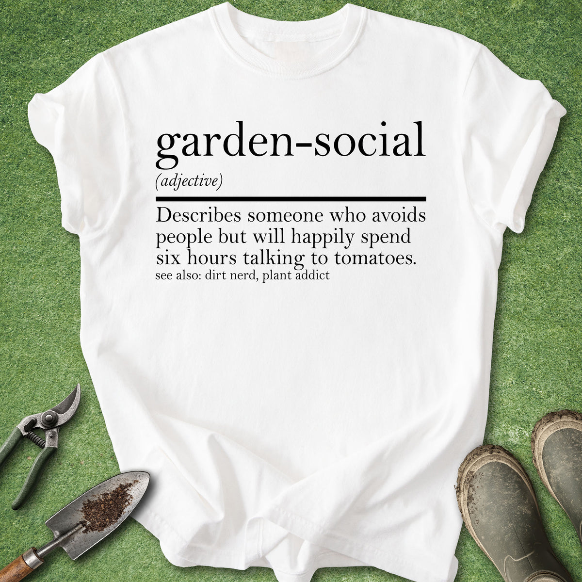 A white graphic T-shirt laid flat on green grass-like background, with printed black text defining the phrase 'garden-social.' Gardening props including pruning shears, a small trowel, and work boots surround the shirt.