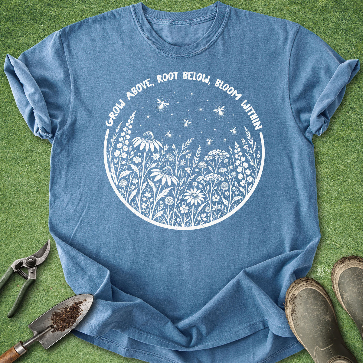 A blue crewneck t-shirt with a white circular graphic of wildflowers, stars, and dragonflies, with the text 'GROW ABOVE, ROOT BELOW, BLOOM WITHIN'. The shirt is laid flat on green grass with gardening tools (trowel, pruning shears) and muddy boots nearby.