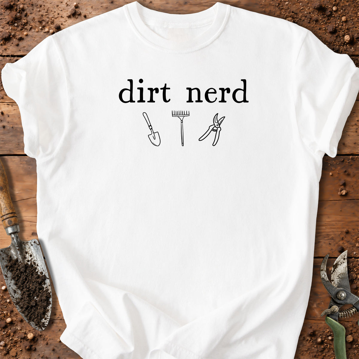 A white short-sleeve t-shirt with a black graphic that reads 'dirt nerd' above simple line drawings of a shovel, rake, and pruning shears. The shirt is laid flat on a rustic wooden surface surrounded by gardening tools and soil.