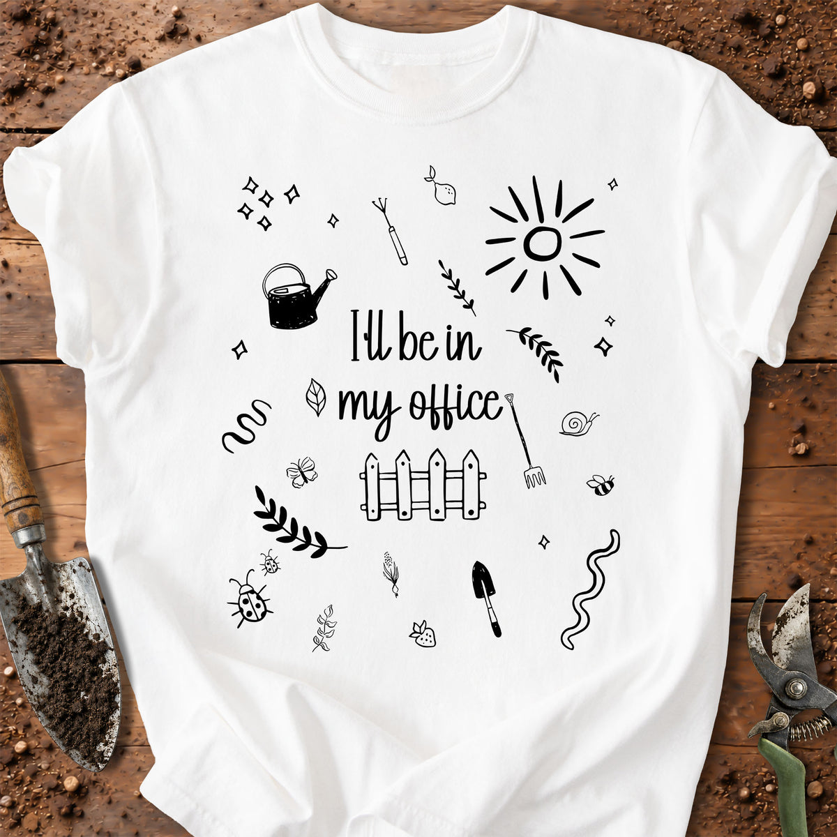 A white short-sleeve crewneck t-shirt with a black hand-drawn garden-themed graphic and the text 'I'll be in my office'. The shirt is displayed in a flat-lay arrangement on a rustic wooden background with gardening props like a trowel, soil, and pruning shears.