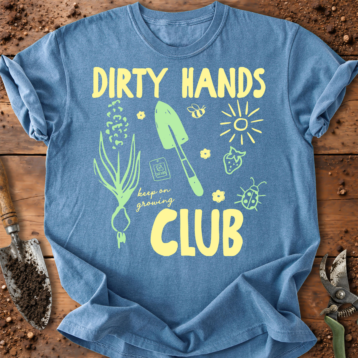 A blue graphic t-shirt laid flat on a rustic wooden surface with gardening props like soil, a trowel, and pruning shears. The shirt features a yellow and green graphic design with the text 'DIRTY HANDS CLUB' and gardening illustrations.