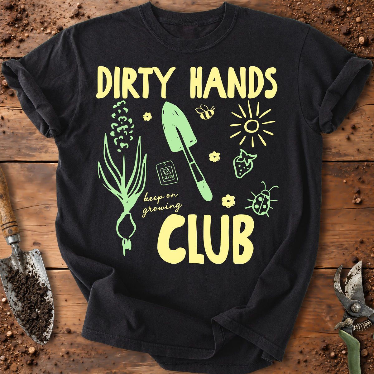 A black graphic t-shirt laid flat on a rustic wooden surface with gardening props. The shirt features yellow text reading 'DIRTY HANDS CLUB' and light green illustrations of a leafy plant, trowel, bee, sun, strawberry, ladybug, and the phrase 'keep on growing'.